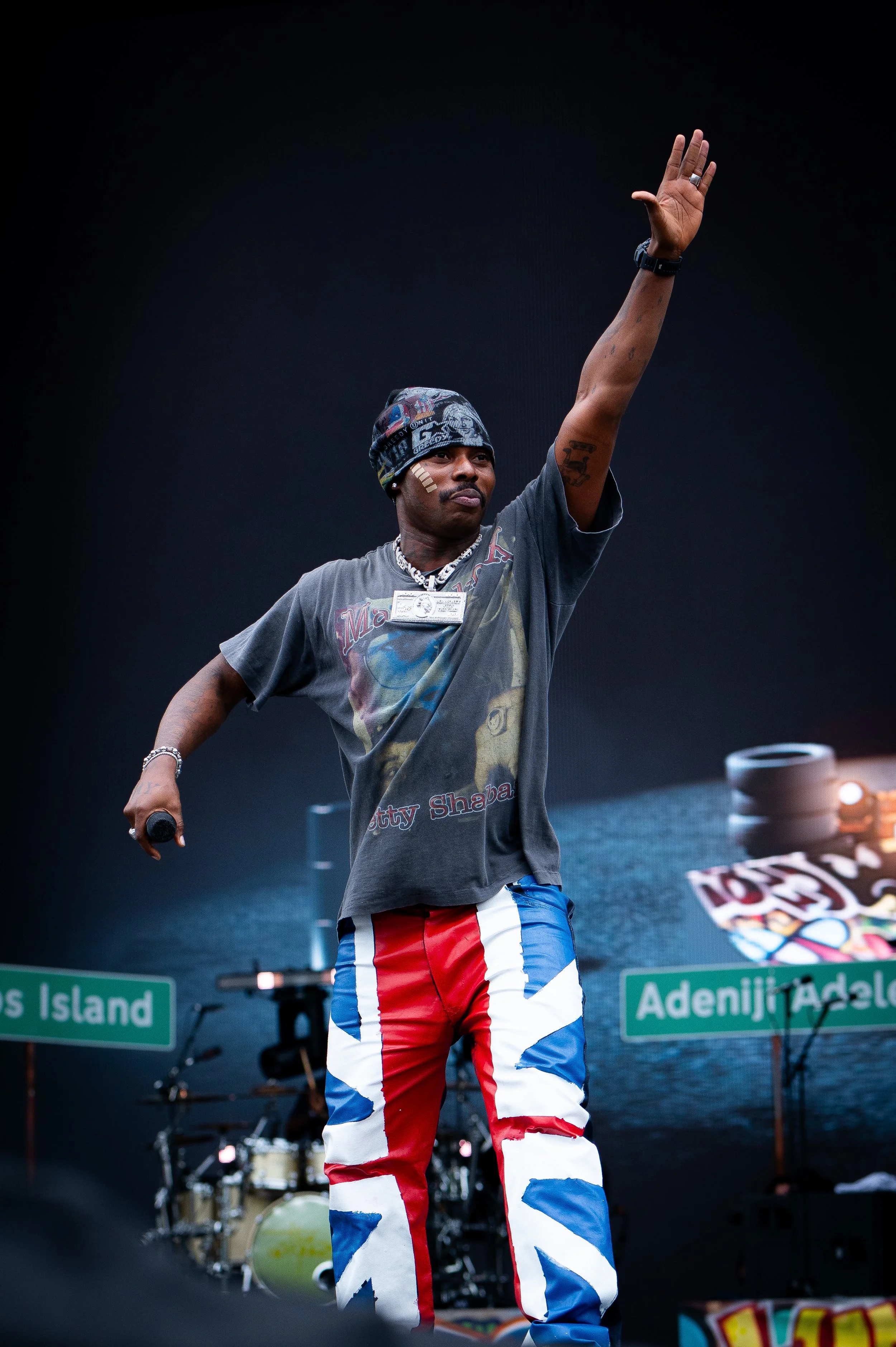 A man dressed in a casual t-shirt and patriotic red, white, and blue pants is on stage, raising one arm with his hand open. He wears jewelry and a hat, and has tattoos on his arm. The background includes microphones, musical instruments, and a sign w