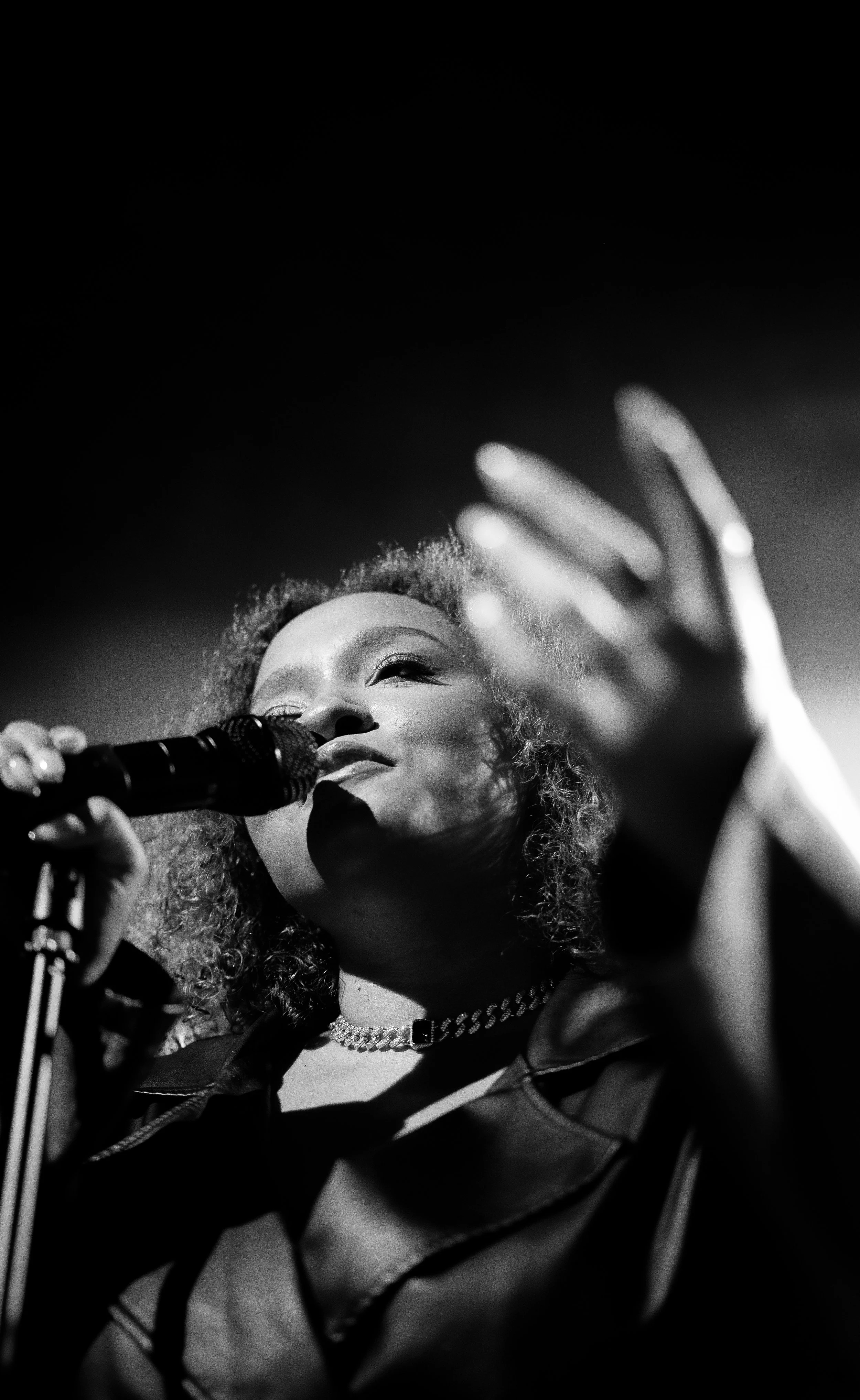 Black and white photo of a woman singing into a microphone on stage, with her eyes closed and a necklace around her neck, raised hand, curly hair, and a confident expression.