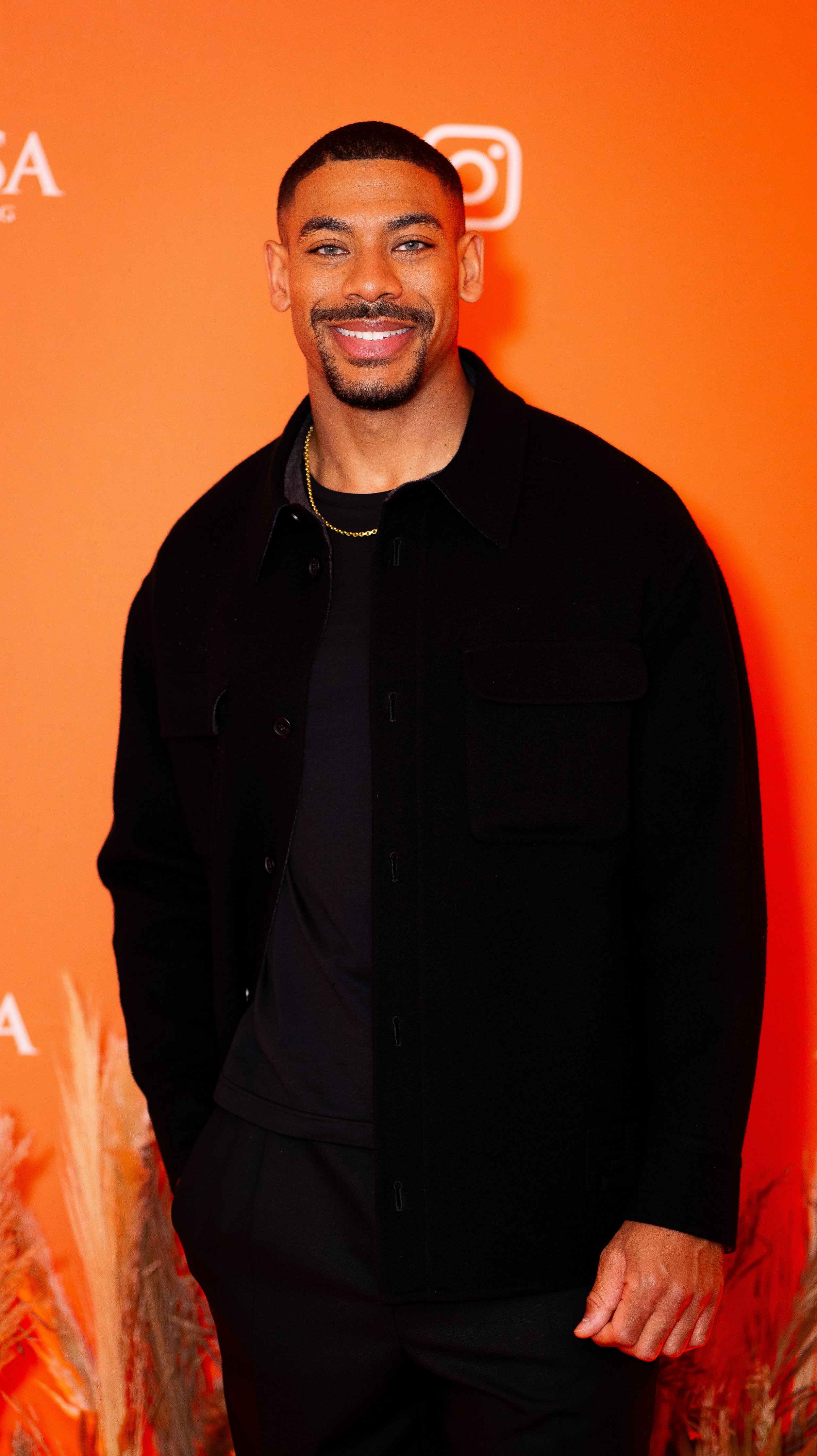 A smiling man with a short haircut and goatee, wearing a black jacket and gold chain, standing against an orange background with social media icons.