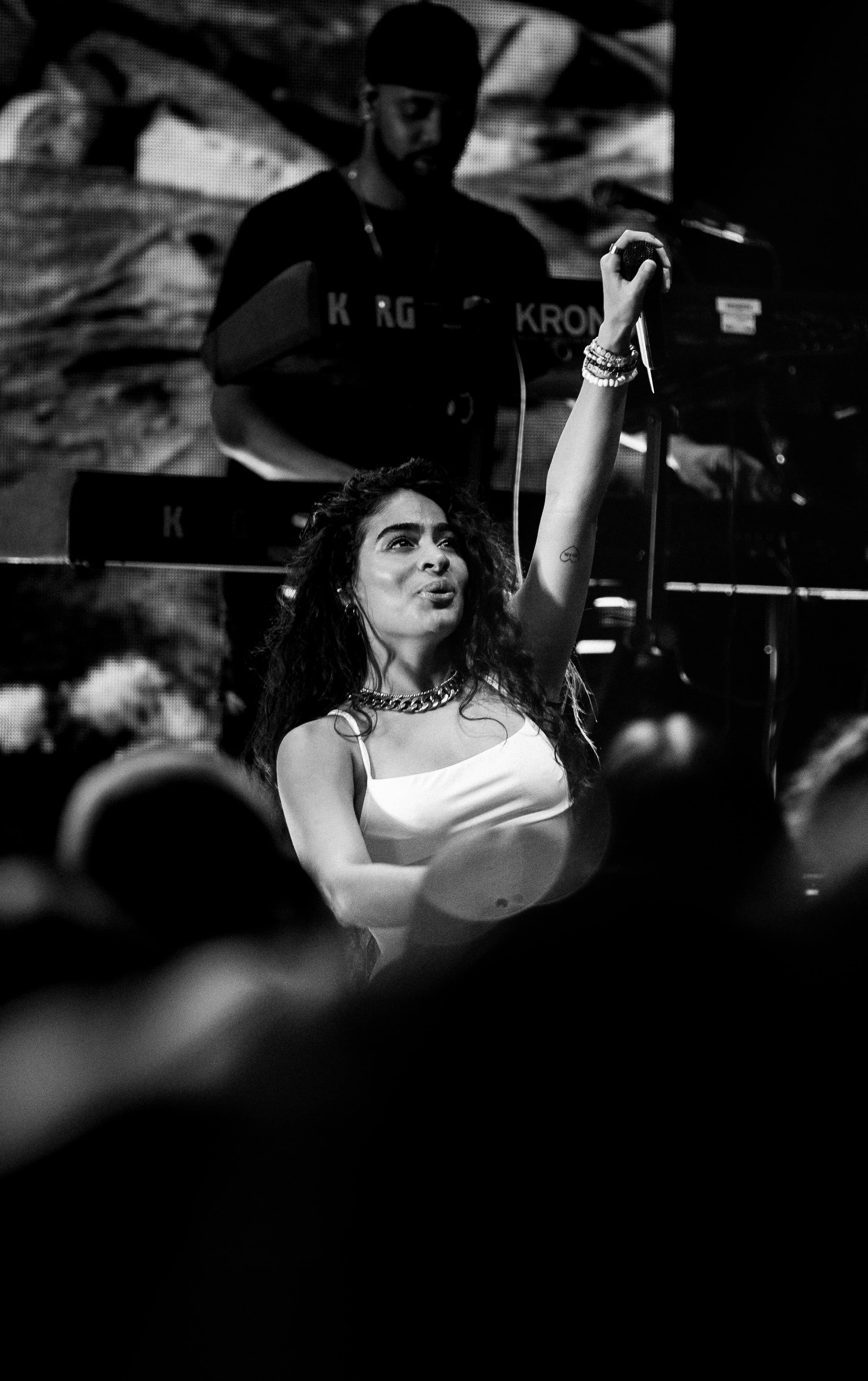 A woman with curly hair taking a selfie with her phone, wearing a white top and multiple bracelets, while a man in the background plays a keyboard.