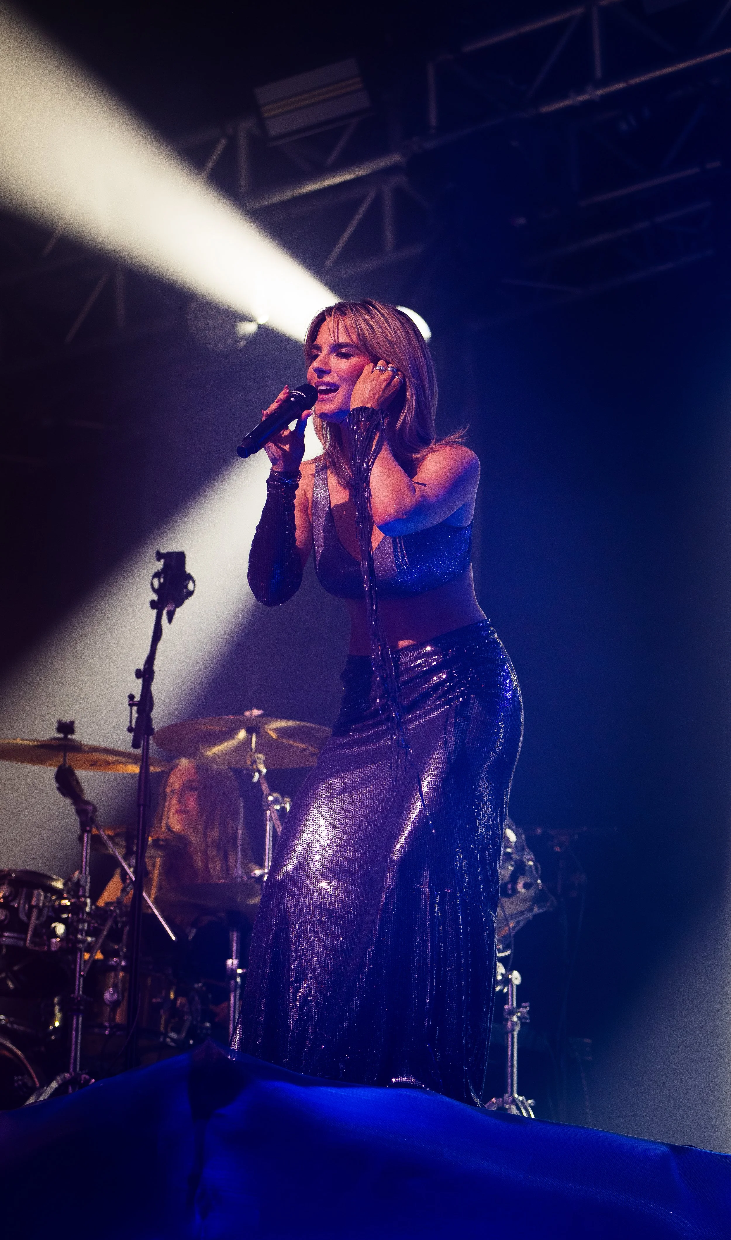 A female singer performing on stage with a microphone, wearing a shiny, metallic purple dress, under stage lights with a band in the background.