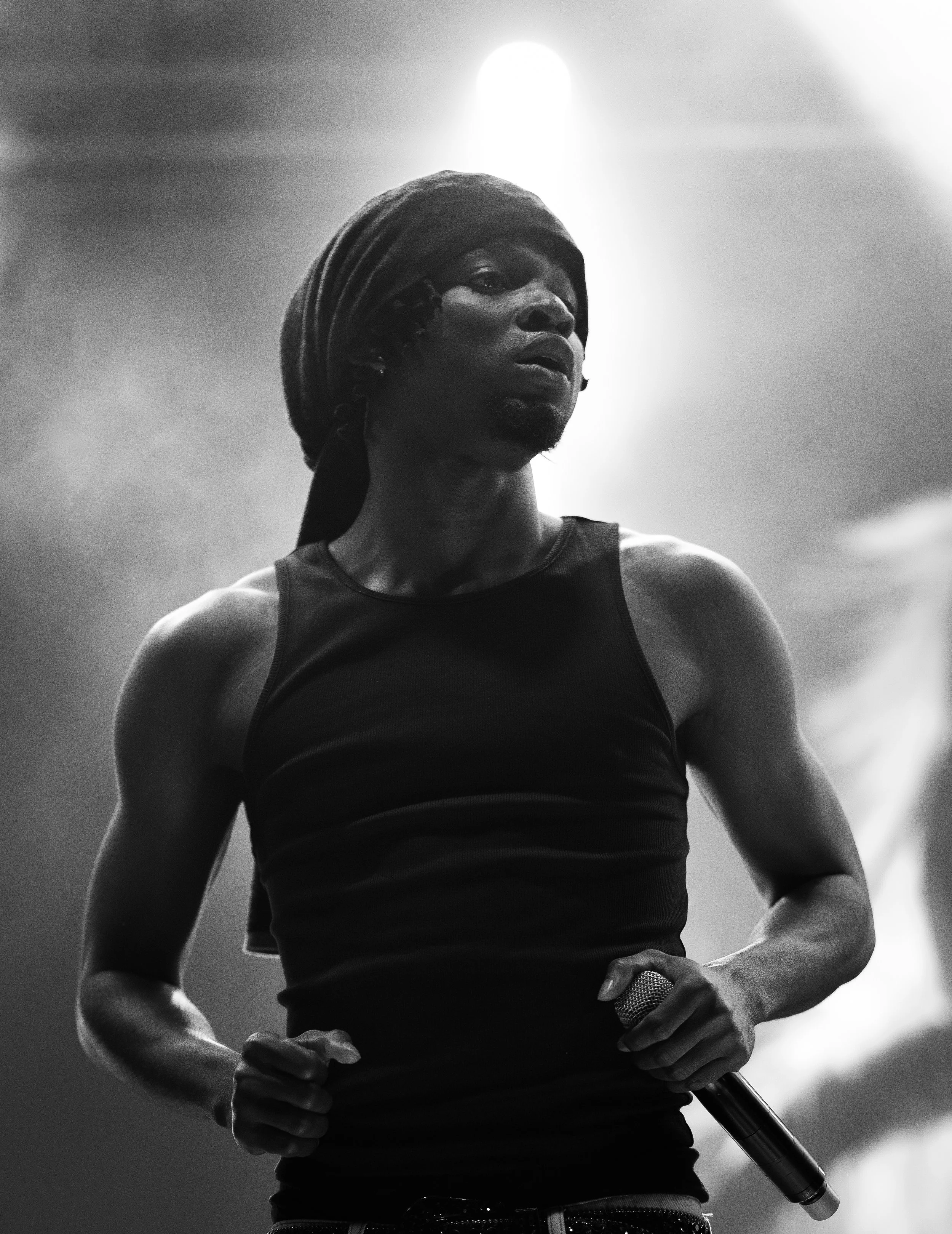 A black and white photo of a woman wearing a headscarf and sleeveless shirt, holding a microphone, with a serious look, in front of a blurry cloudy sky with sunlight behind her.