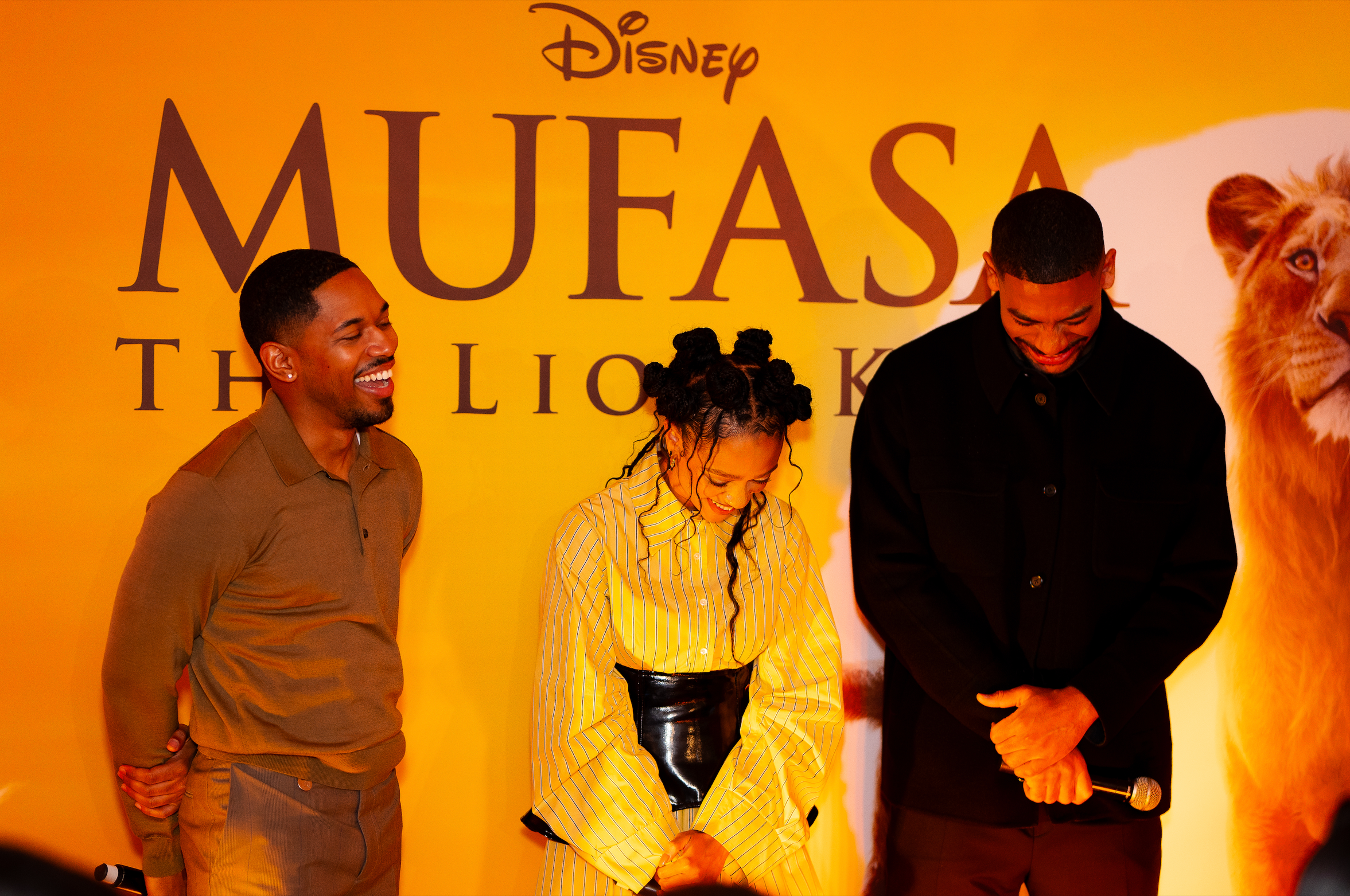 Three people standing and smiling at an event with a yellow backdrop featuring the text 'Disney Mufasa the Lion' and a lion's face on the right side.