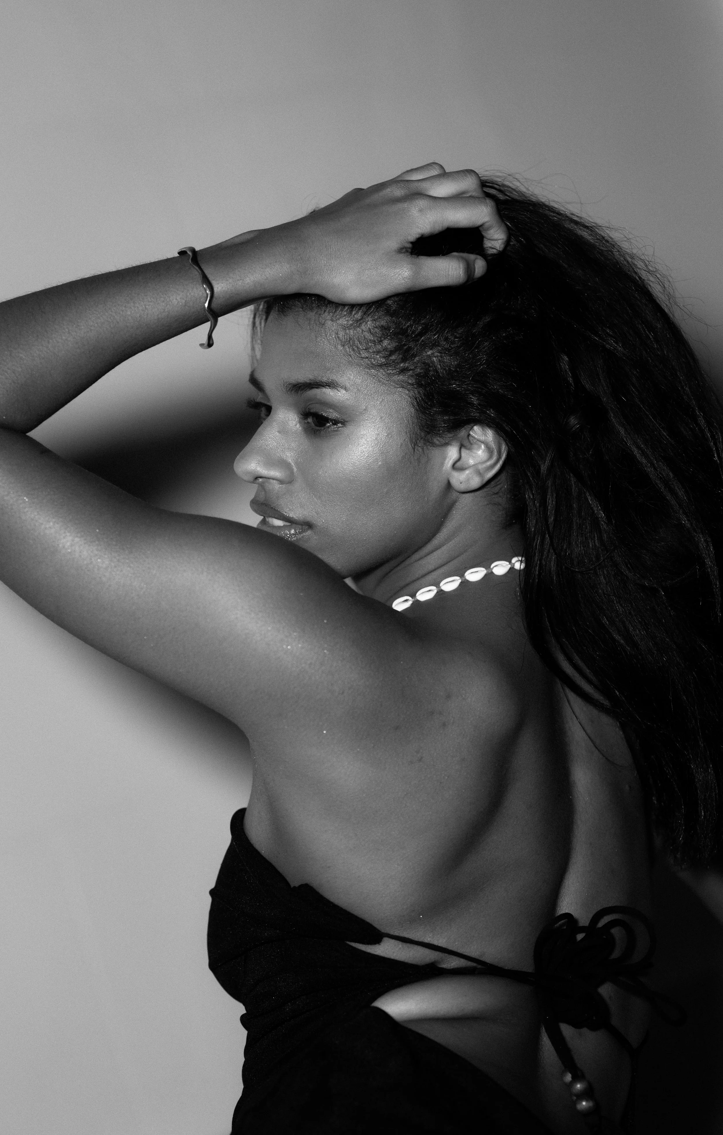 A woman with long dark hair, wearing a pearl necklace and a black strapless top, poses with her right arm raised and her hand on her head, showcasing her profile. The photo is in black and white.
