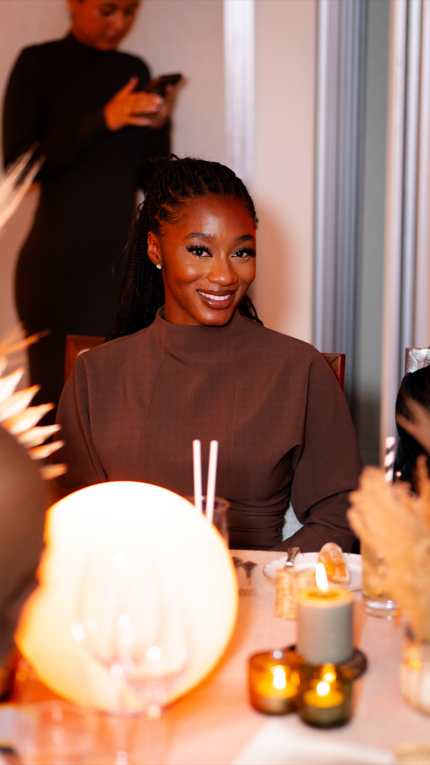 A smiling Black woman with braided hair and earrings sitting at a dinner table, with a woman in black in the background looking at her phone.