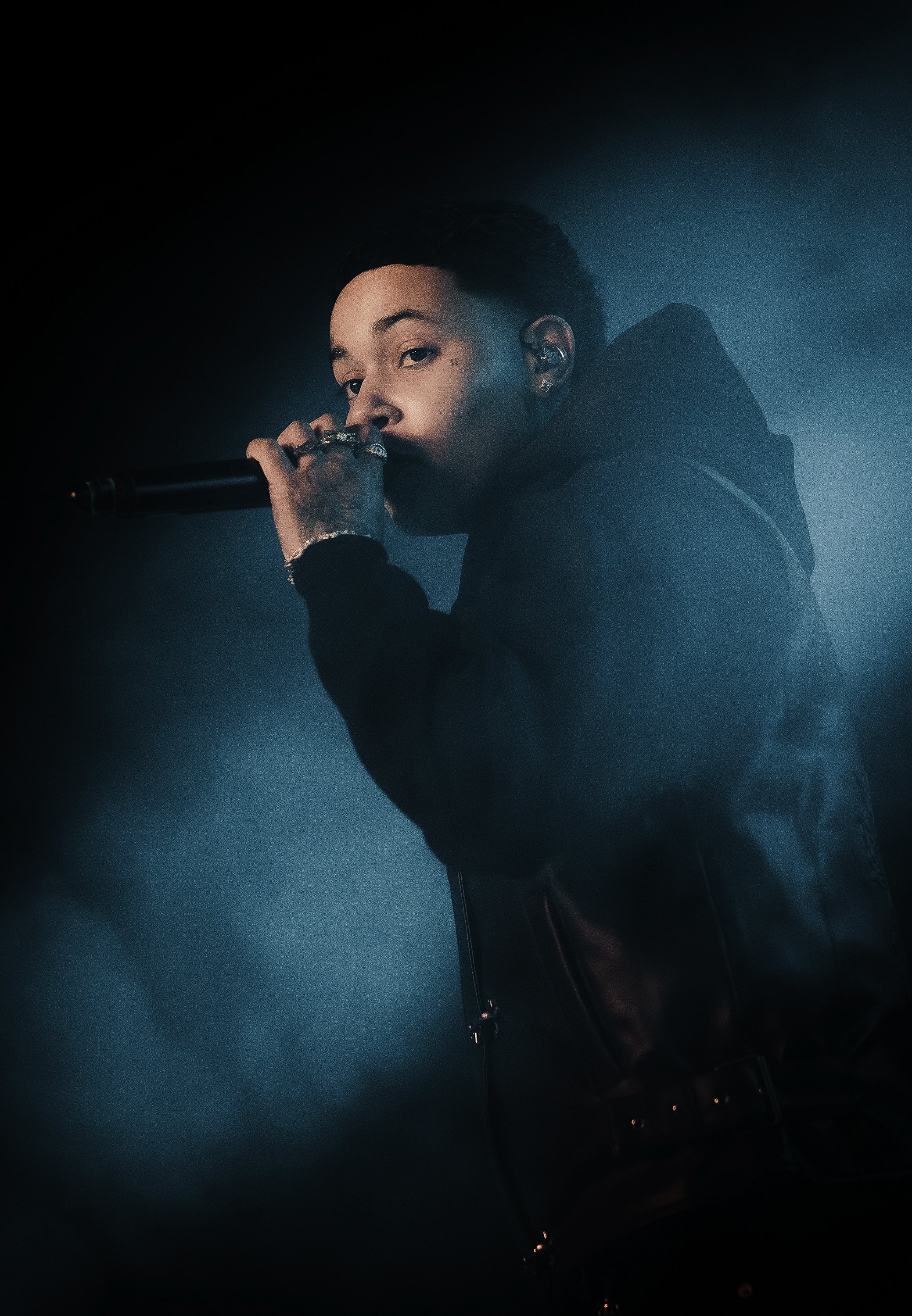 A person holding a microphone close to their mouth, looking over their shoulder, with dim blue lighting and a dark background.