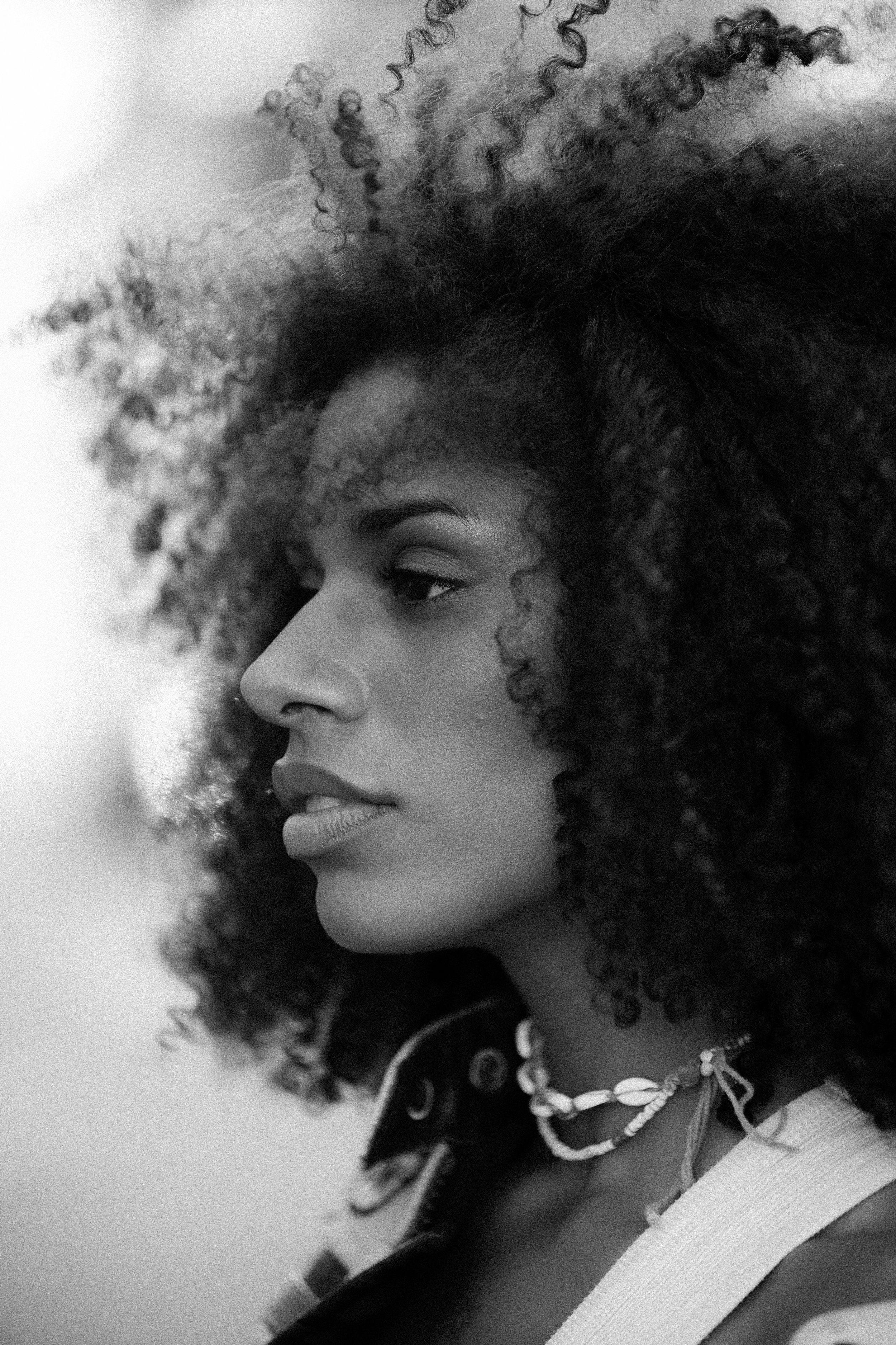 A black and white close-up of a woman's face in profile with curly hair, wearing a patterned top, necklace, and focused expression.