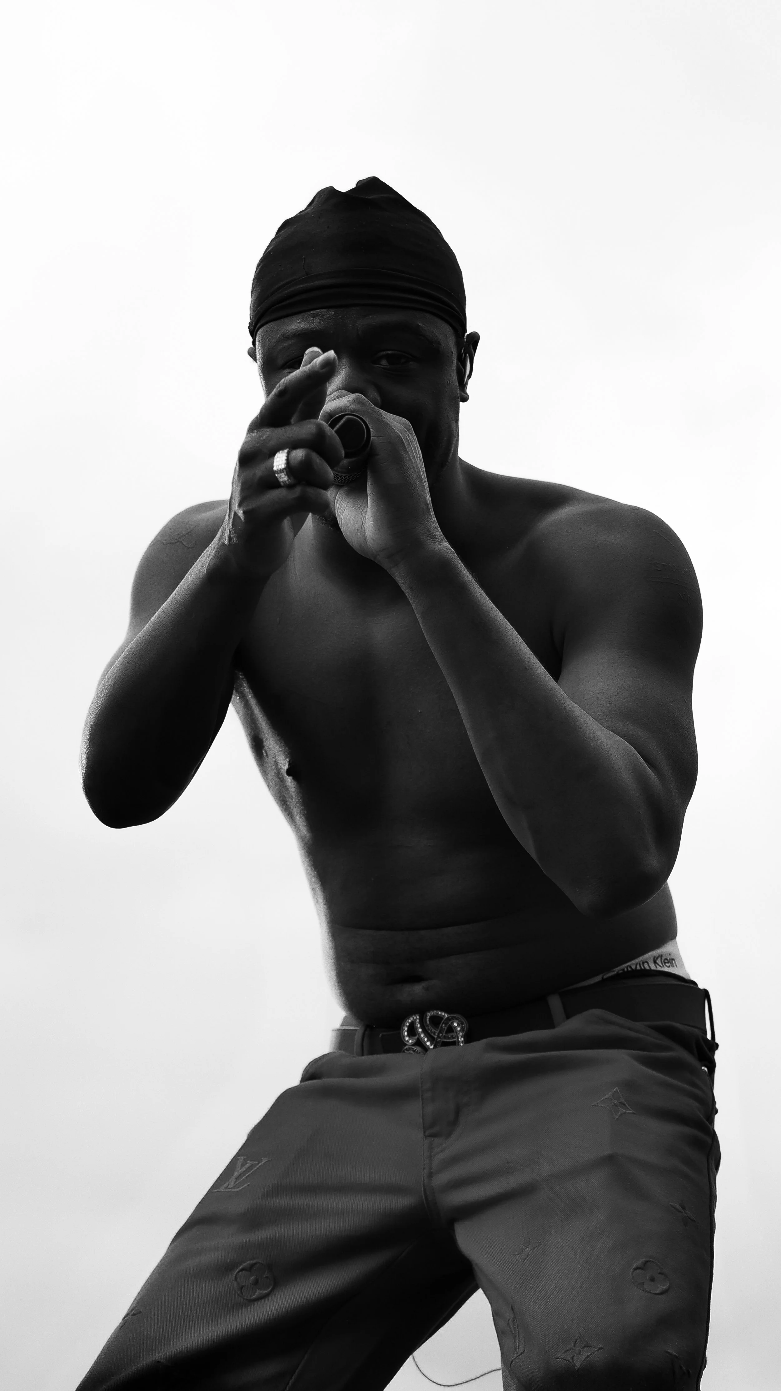 A shirtless man wearing a black head covering and dark pants with logo patterns, holding a camera or similar device up to his face, with a minimalistic, featureless background.