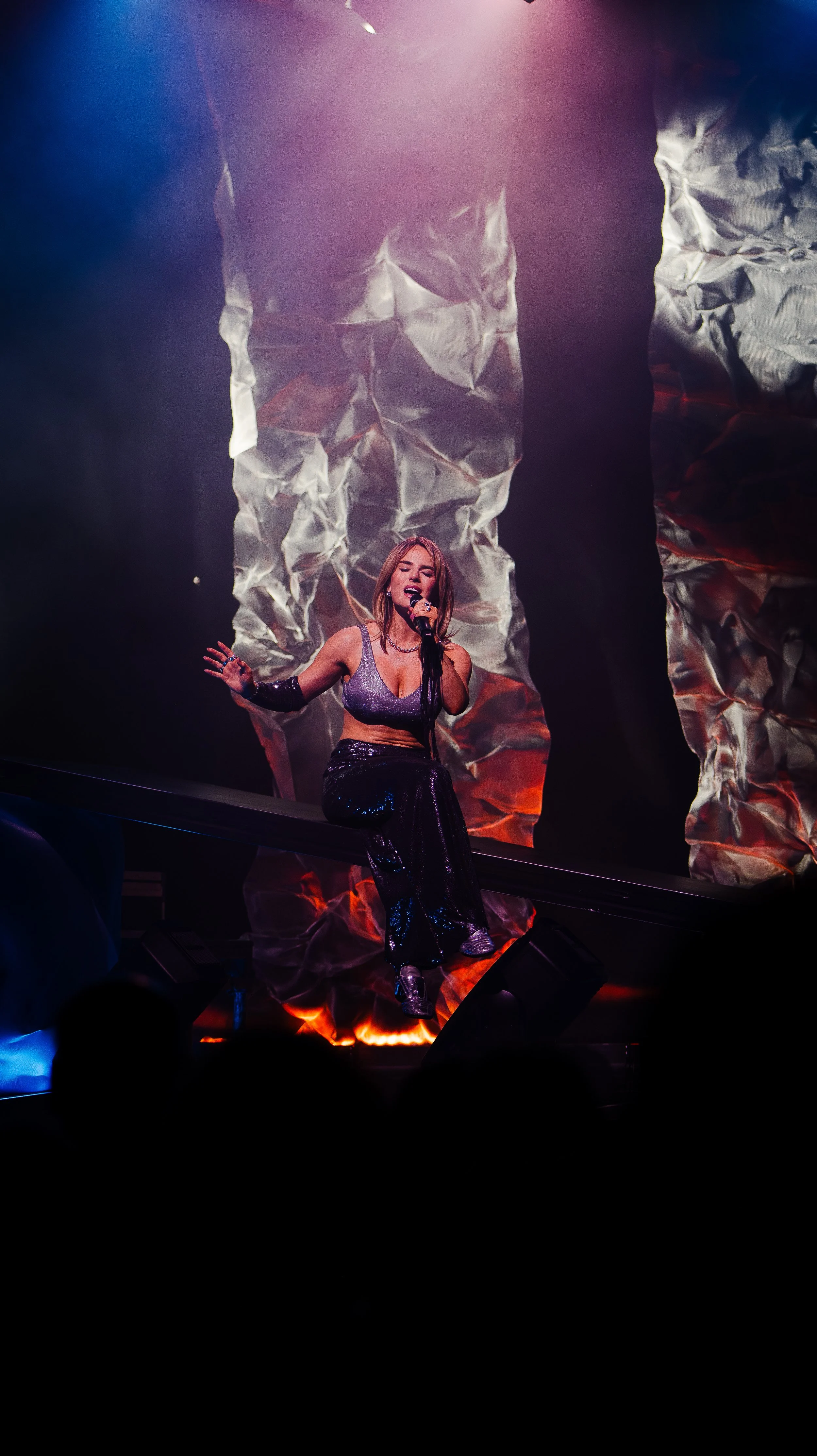 A female singer performing on stage with a microphone, dressed in a shiny crop top and shiny pants, with crumpled metallic backdrops and stage lighting creating a dramatic atmosphere.