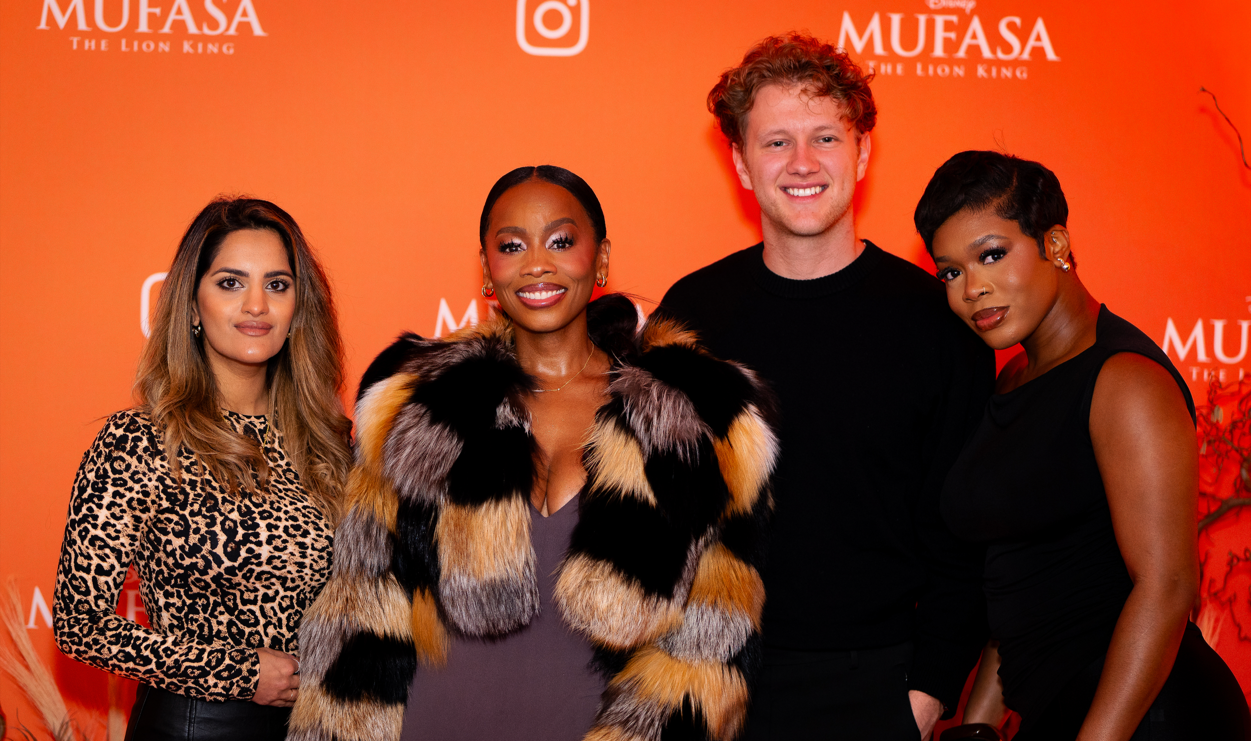Group of four women and one man standing together at an event with an orange background. The women are wearing fashionable outfits, including animal print and fur. The background features the text 'MUFASA The Lion King' and social media icons.