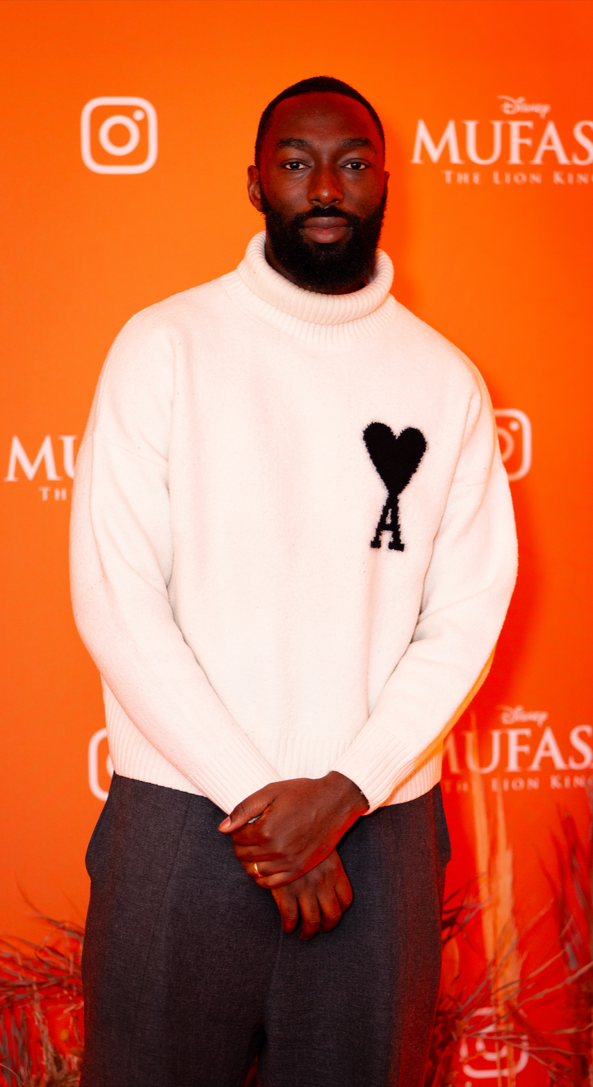 A man standing in front of an orange backdrop with Disney's "The Lion King" and "Mufasa" logo, wearing a white turtleneck sweater with a black heart and letter 'A' on it, and dark pants.