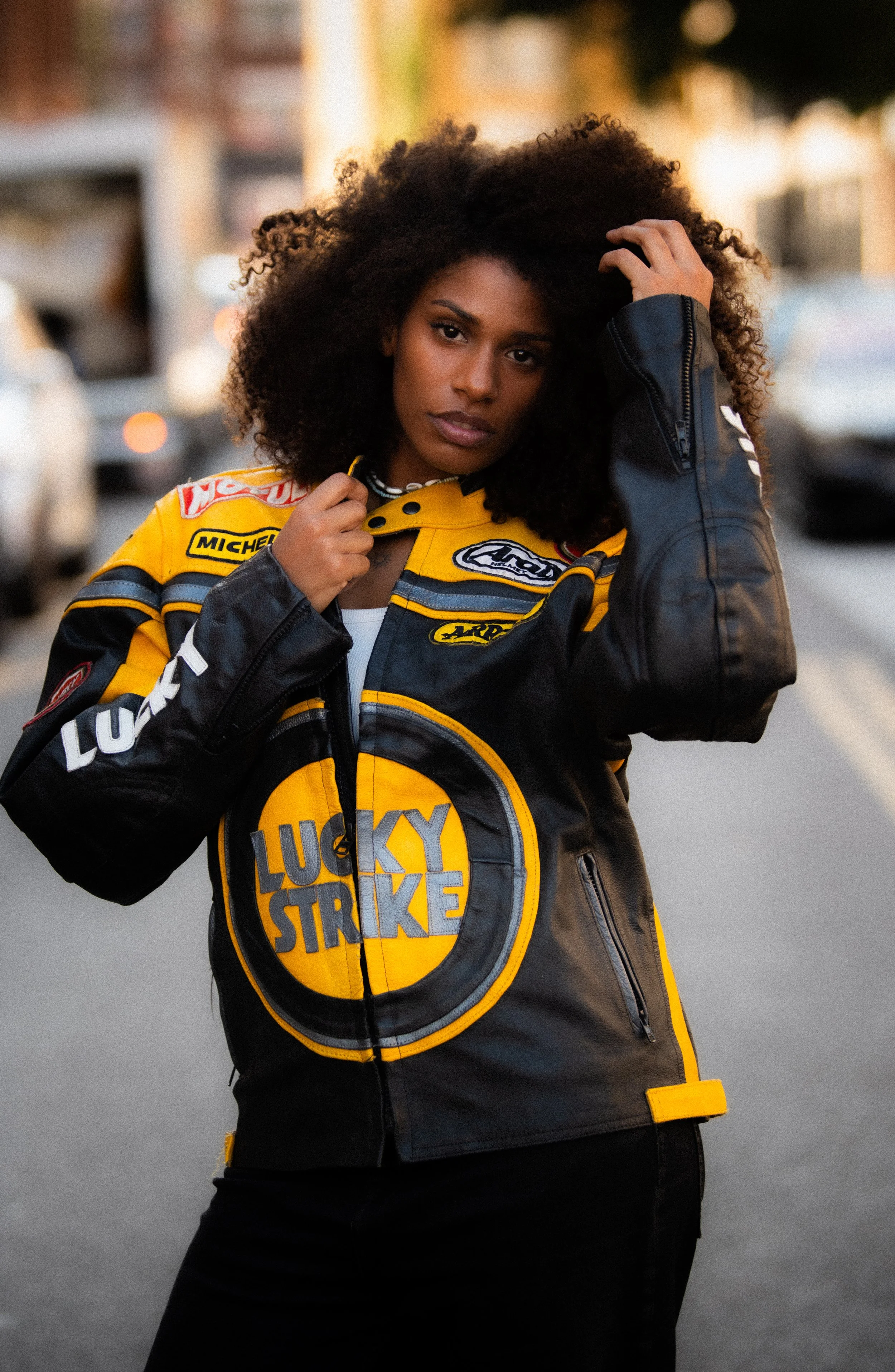 A woman with curly black hair wears a Racing World motorcycle racing jacket with a large Patch that says 'Lucky Strike' on the front, standing outdoors with blurred buildings and cars in the background.