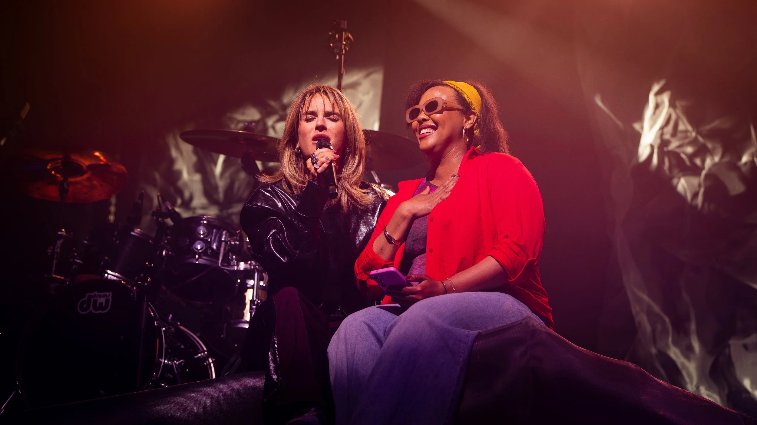 Two women performing on stage under warm lighting, one singing into a microphone and the other smiling with sunglasses and a yellow headband, seated with a phone in hand, with a drum set in the background.