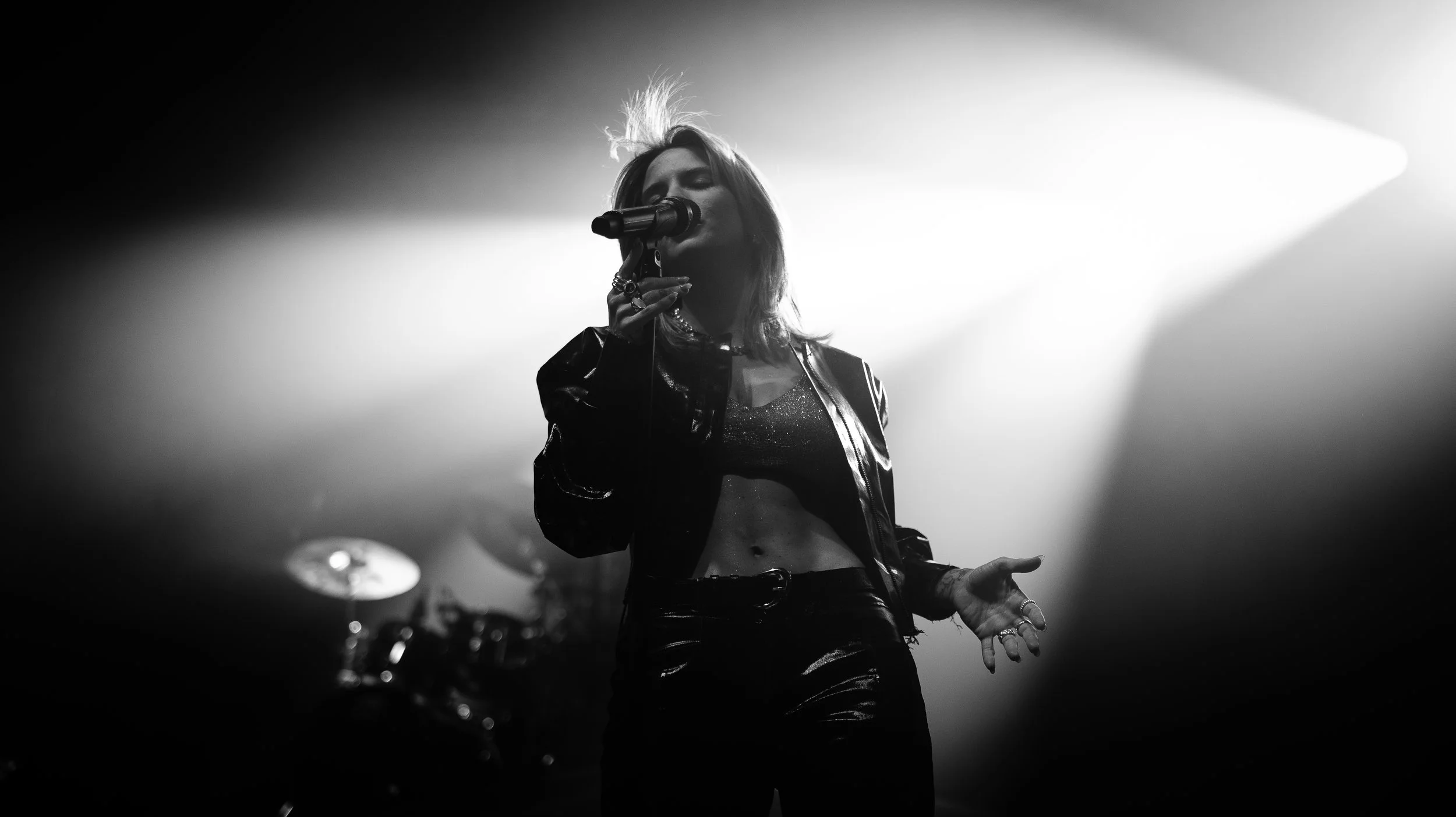 A female singer performing on stage with a microphone, wearing a shiny jacket and crop top, with stage lights creating a dramatic background.