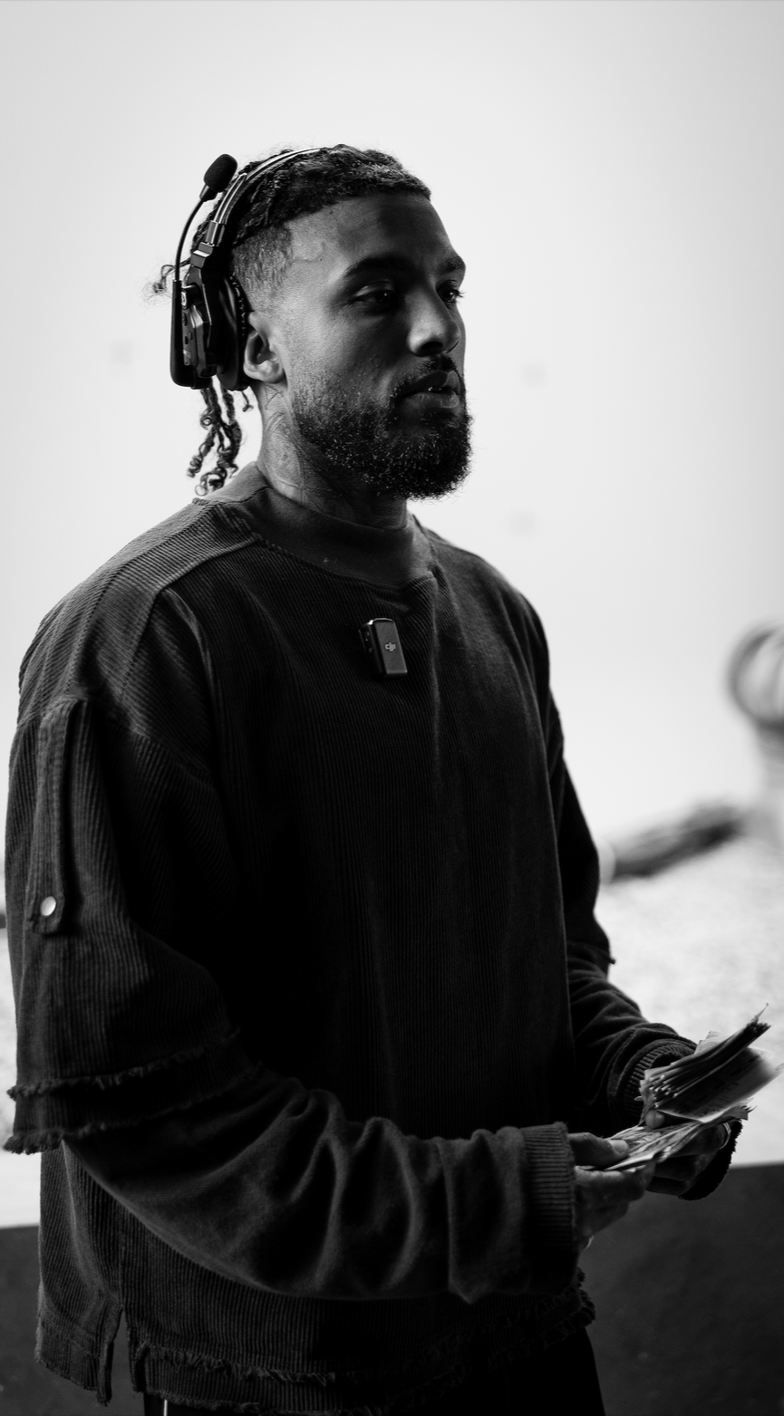A man wearing headphones and a dark sweatshirt, holding papers, standing indoors with a plain wall background.