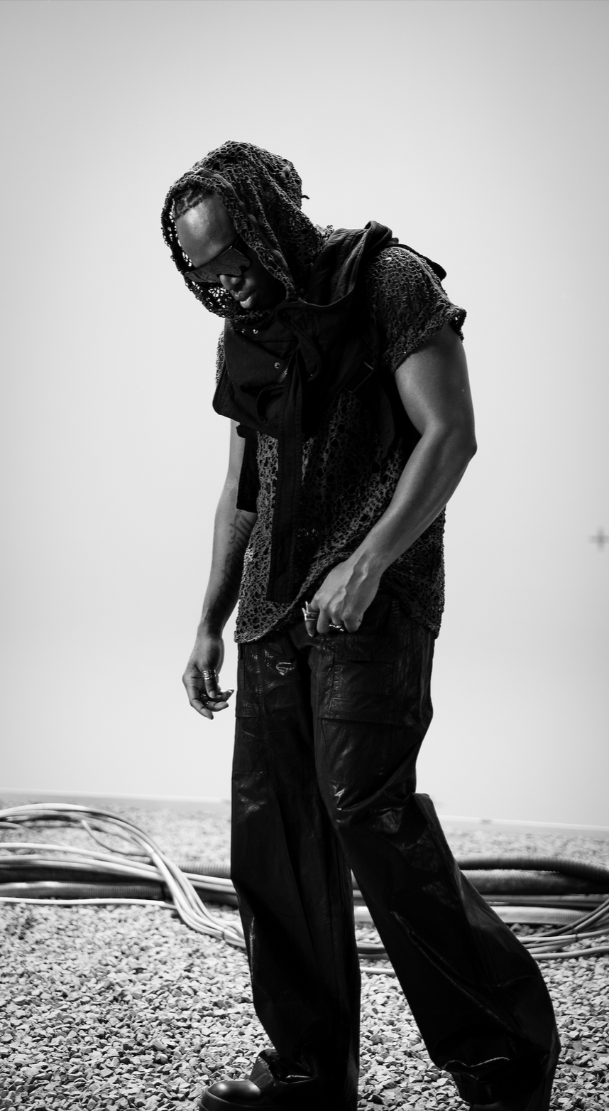 A man with dreadlocks wearing sunglasses, a textured shirt, a vest, and baggy pants, standing on gravel with cables on the ground, looking down.