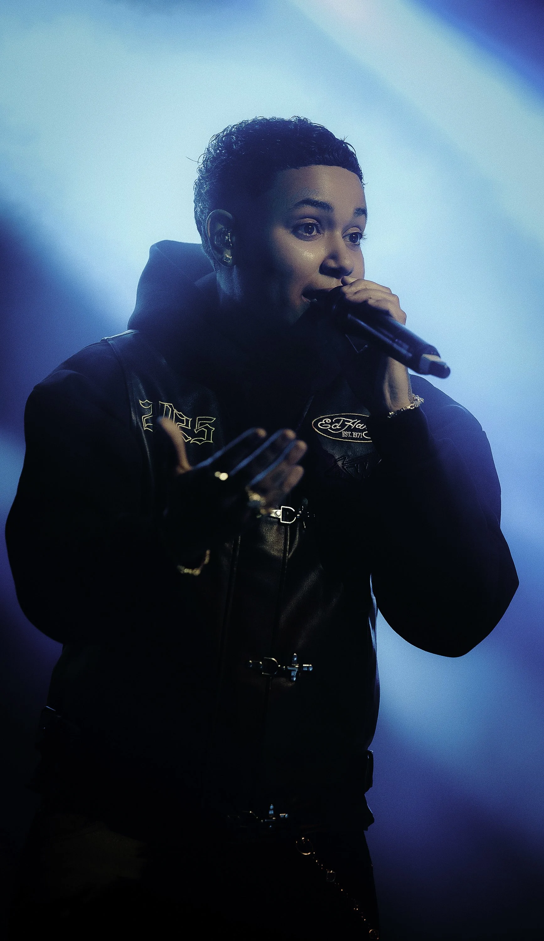 A woman with short curly hair singing into a microphone, wearing a black jacket with patches and jewelry, against a blue gradient background.