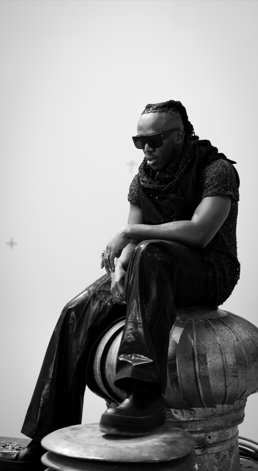 A black-and-white photo of a man sitting on a large, industrial-looking metal object against a plain light background. He wears dark sunglasses, a short-sleeved textured shirt with a scarf, shiny leather pants, and chunky boots with an oval platform. He has braided hair and several rings on his fingers.