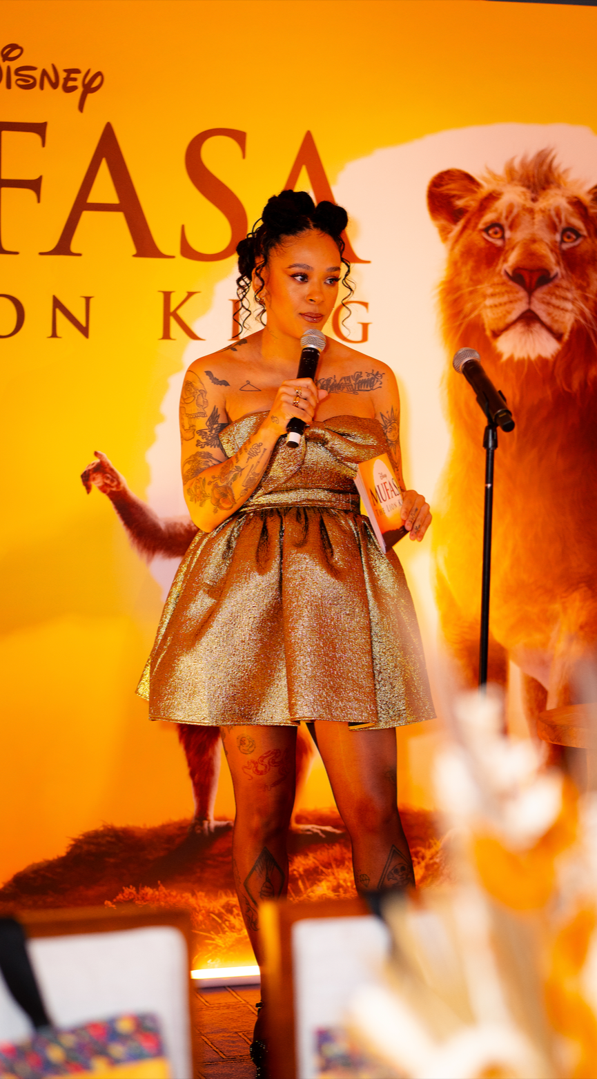 A woman with tattoos on her arms and legs, wearing a shiny gold dress, holding a microphone, standing in front of a yellow background with a lion and a fox, and text that reads "Disney FASA on King."