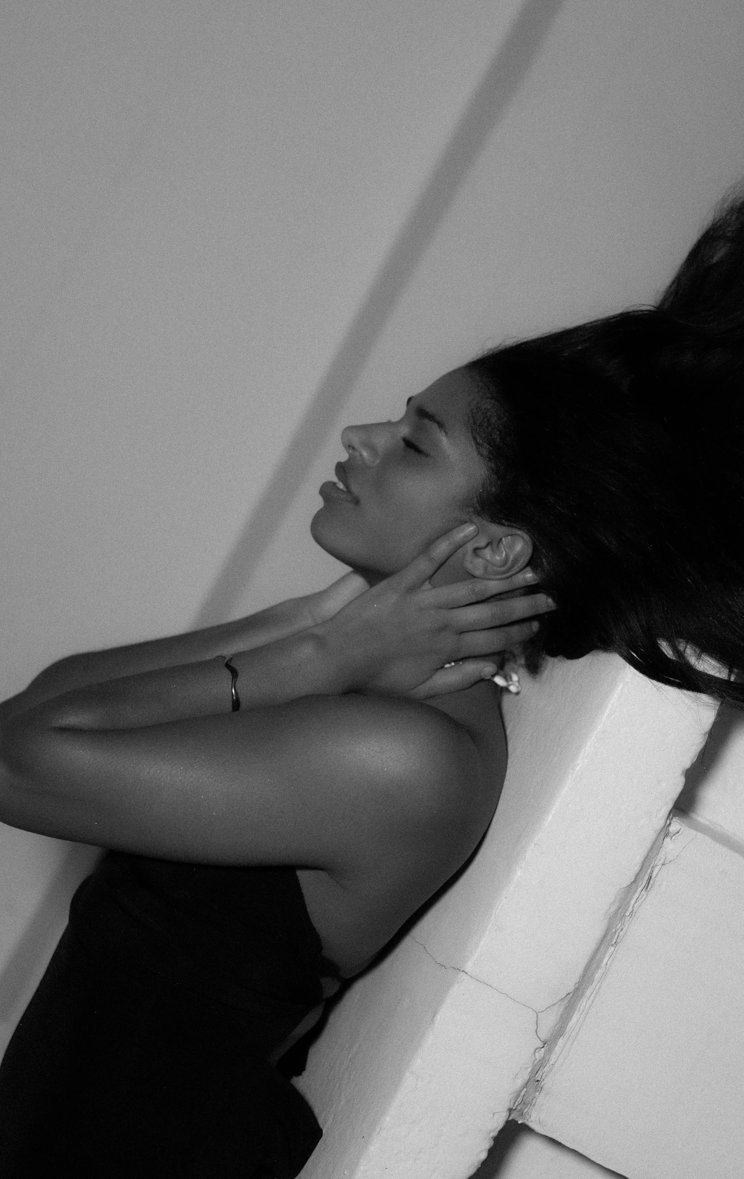 A woman with dark hair and smooth skin leaning against a wall, resting her head on her hand with eyes closed and a slight smile.