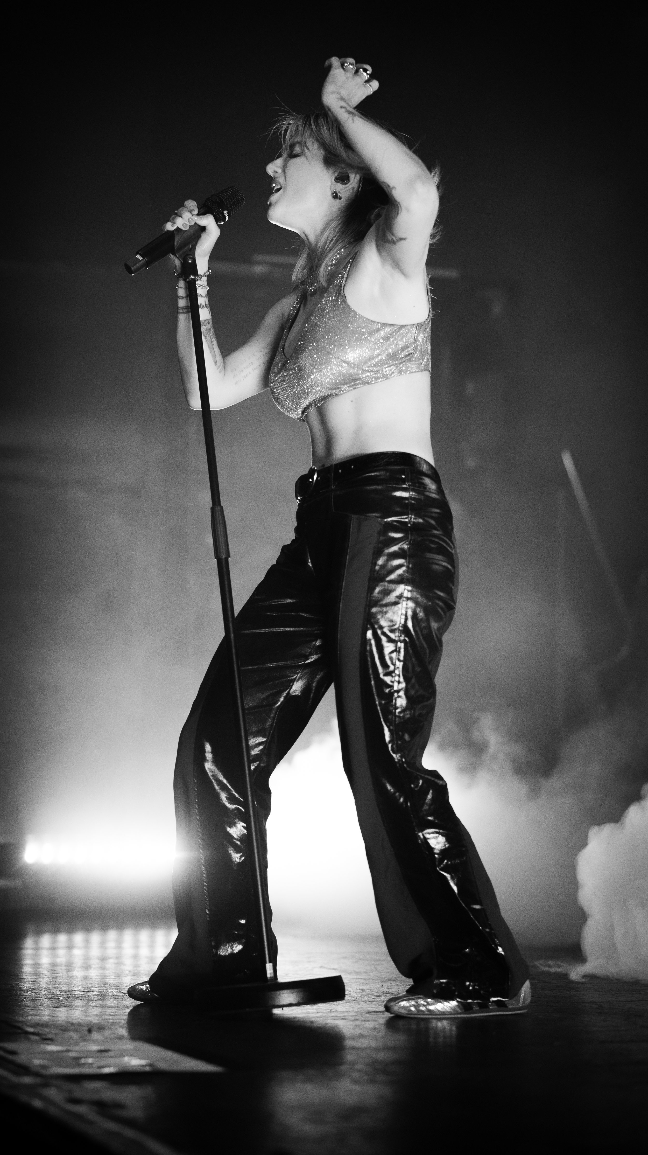 A female singer passionately performing on stage with a microphone, wearing shiny top, leather pants, and metallic shoes, mid-motion with one arm raised and eyes closed, in black and white.