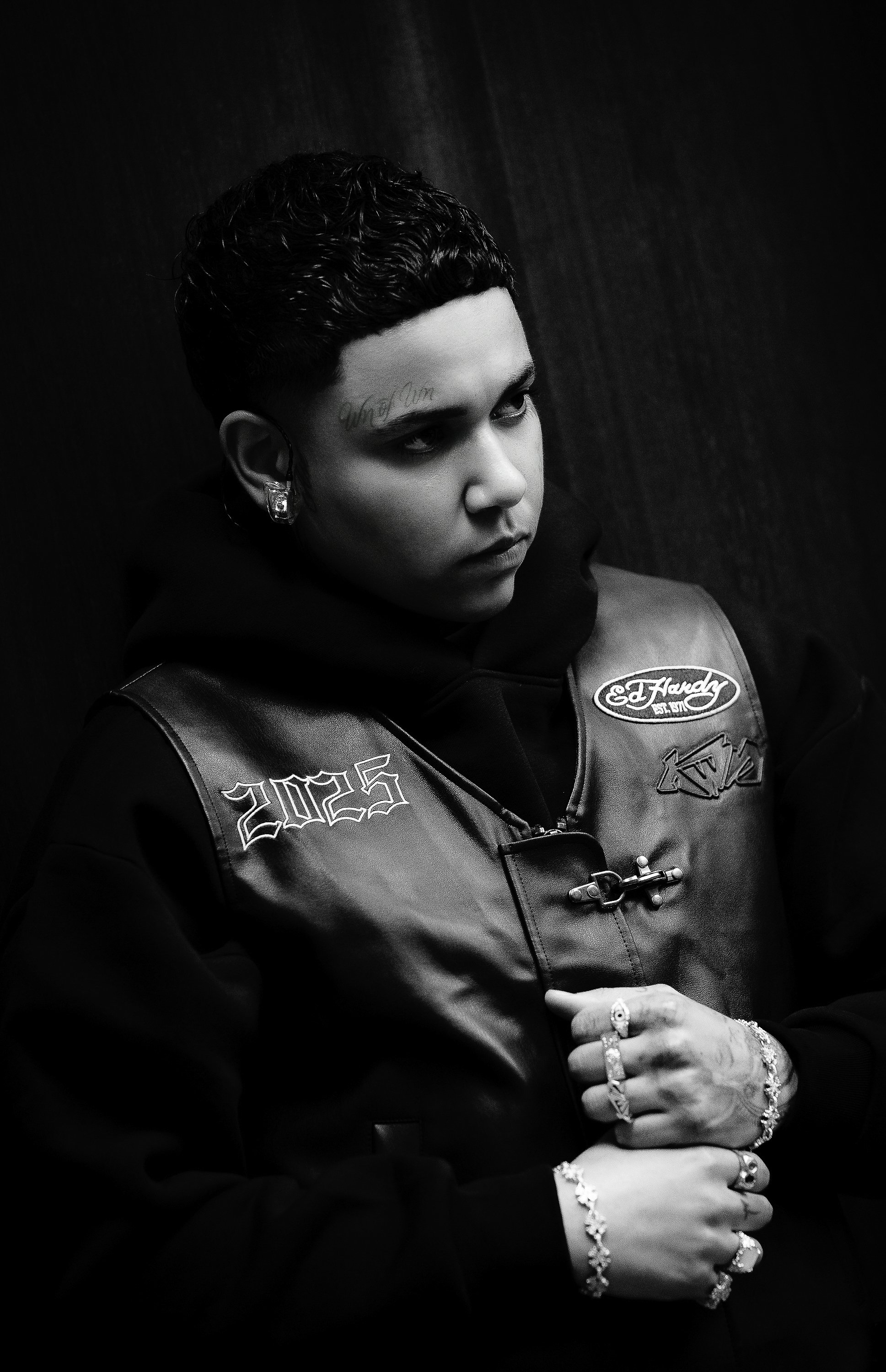 Black and white photo of a woman with short, dark, curly hair, wearing a leather jacket with patches, earrings, rings, and bracelets, looking to the side with a serious expression.