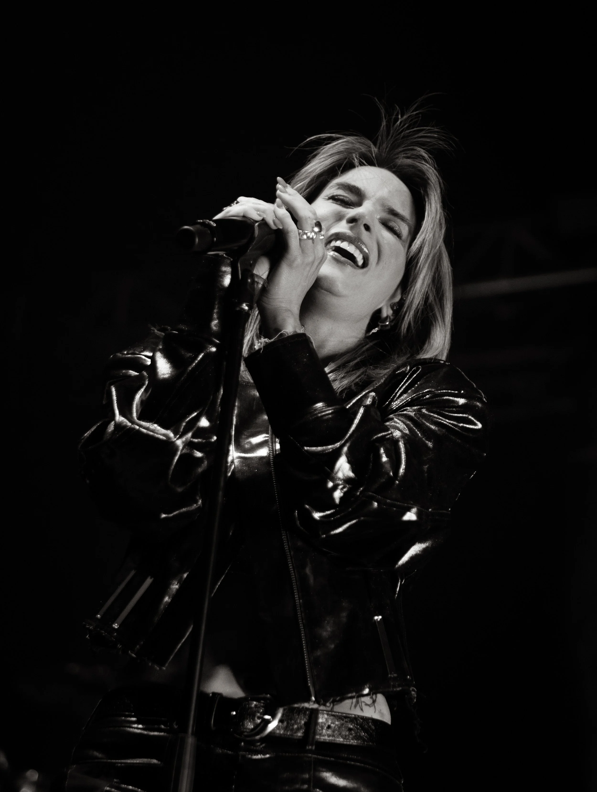 Black and white photo of a woman singing into a microphone, wearing a shiny leather jacket, with her eyes closed and her mouth open.