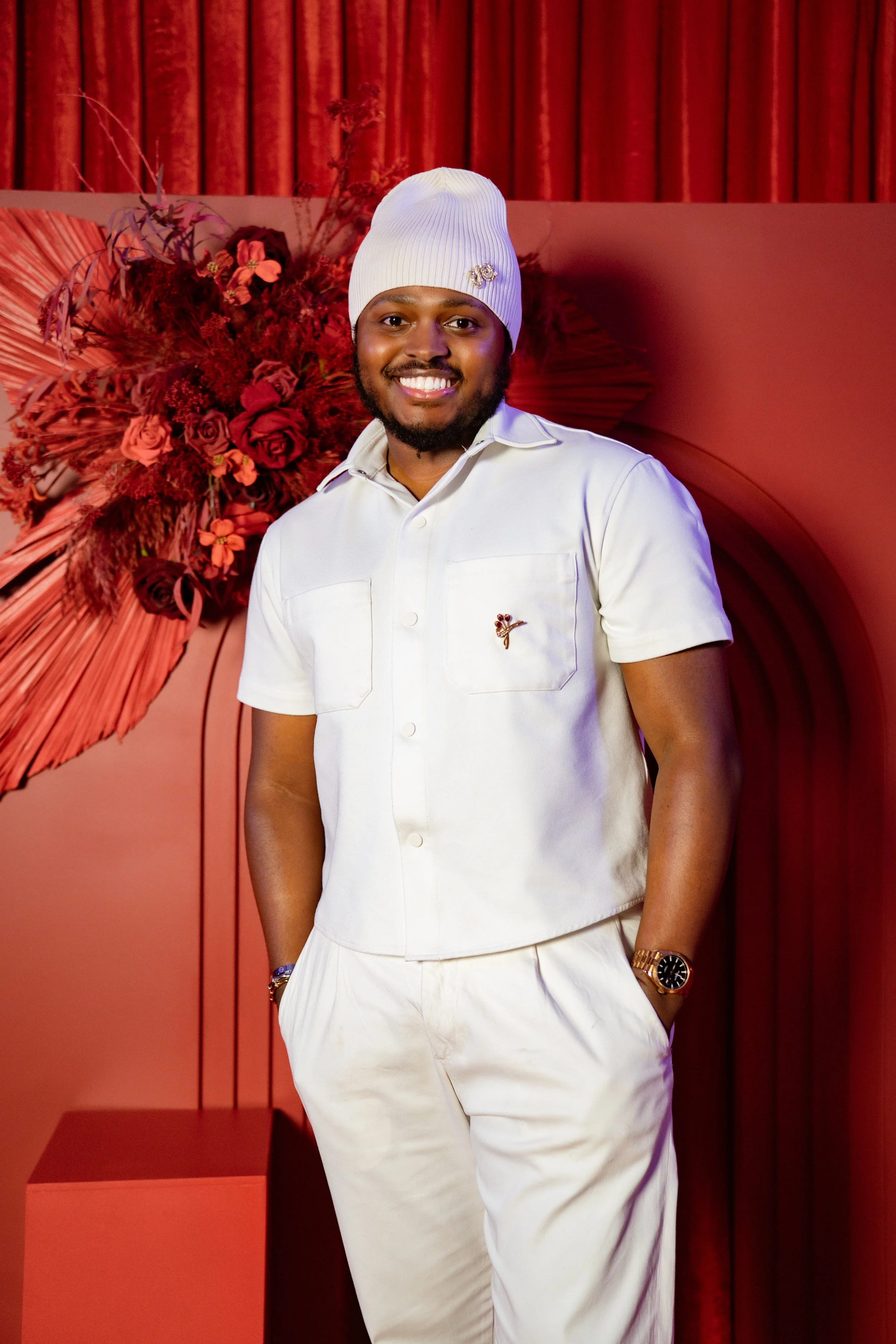 A smiling man wearing a white beanie and a white short-sleeved shirt with a pin, standing in front of a red curtain and floral arrangement, with a red wall and platform in the background.