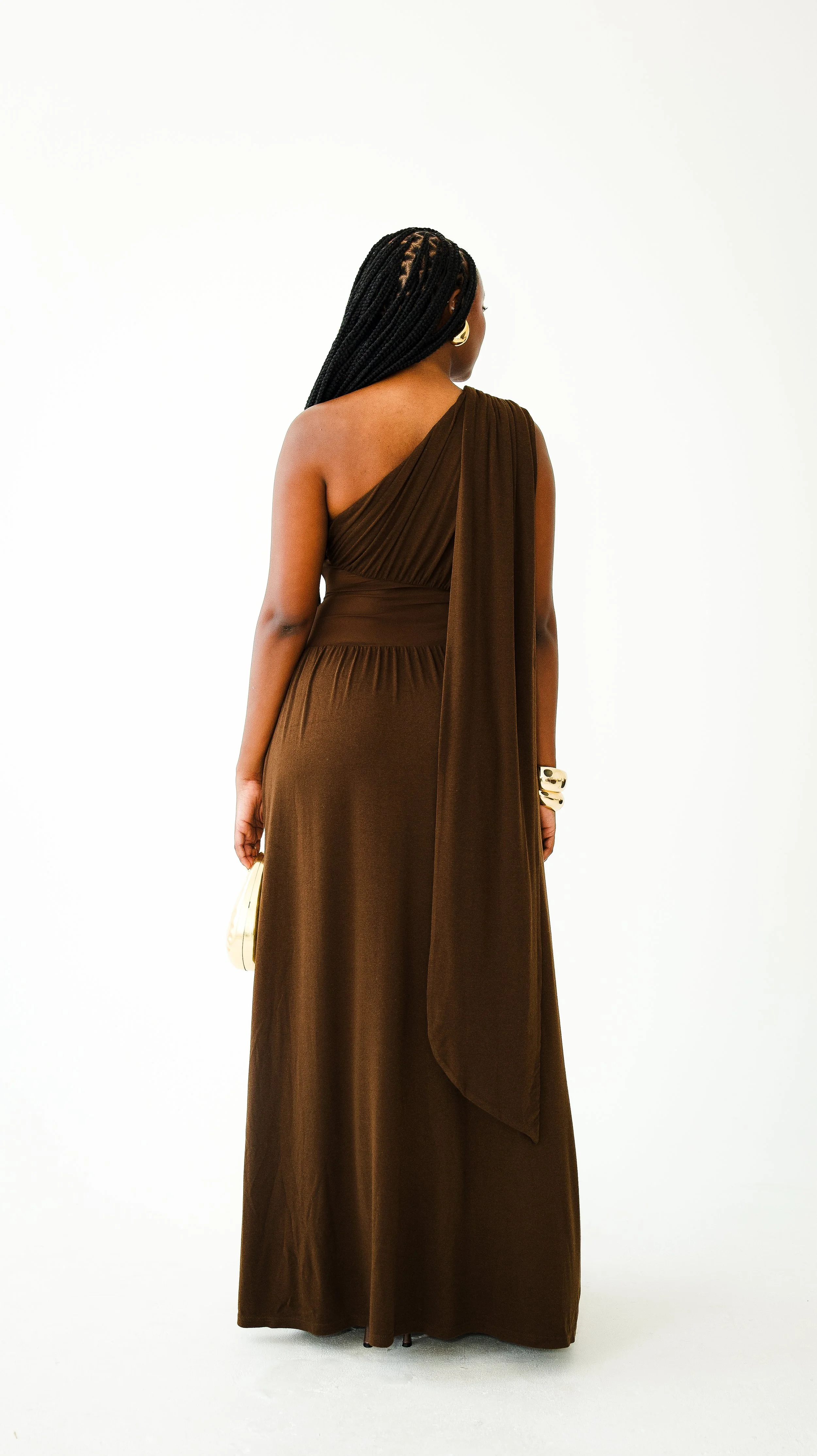 Back view of a woman wearing a long, brown, one-shoulder dress with pleated fabric and large gold earrings, standing against a plain white background.