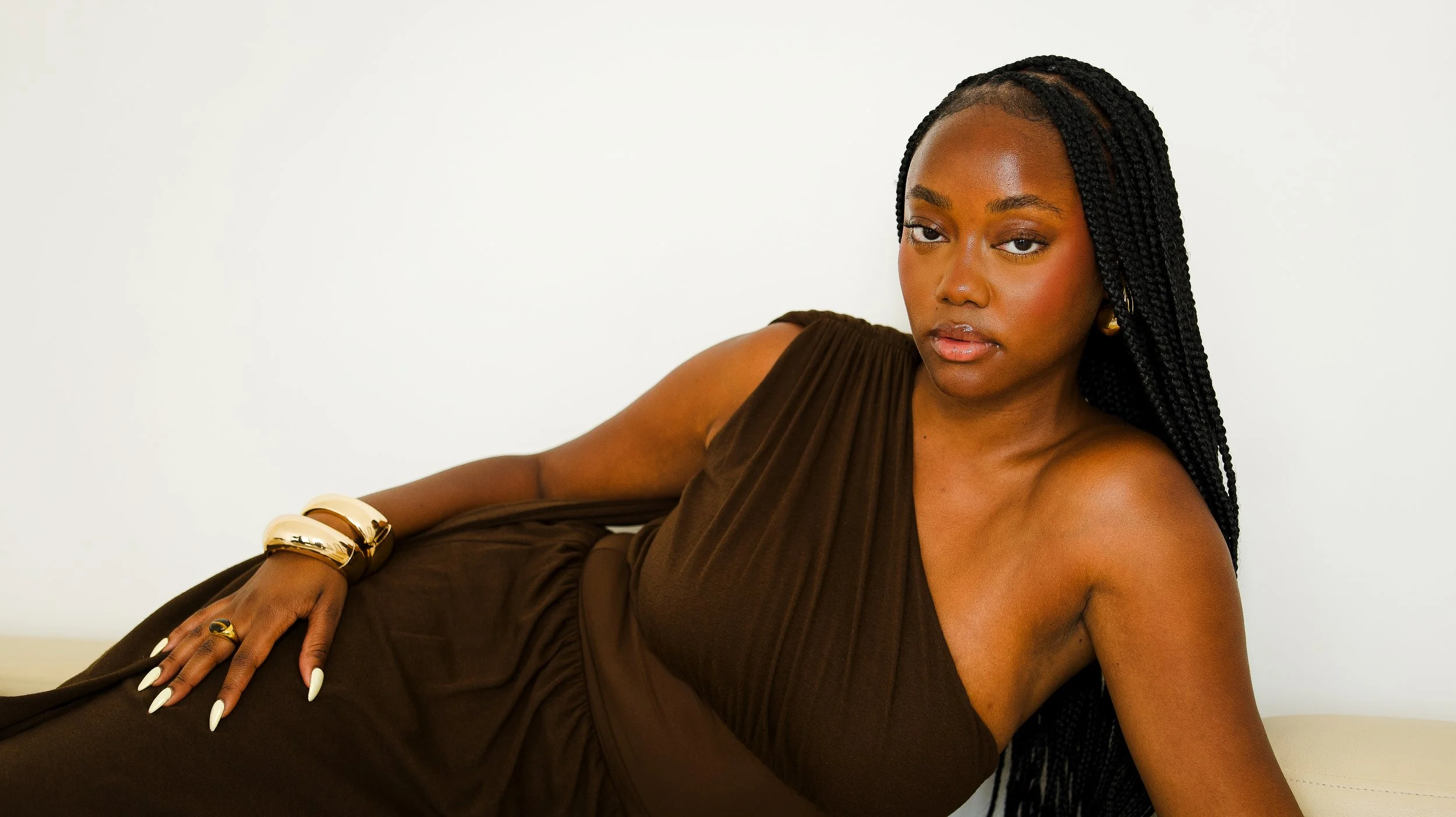A woman with braided hair and brown skin lying on her side against a plain light-colored background, wearing a brown dress and gold jewelry.
