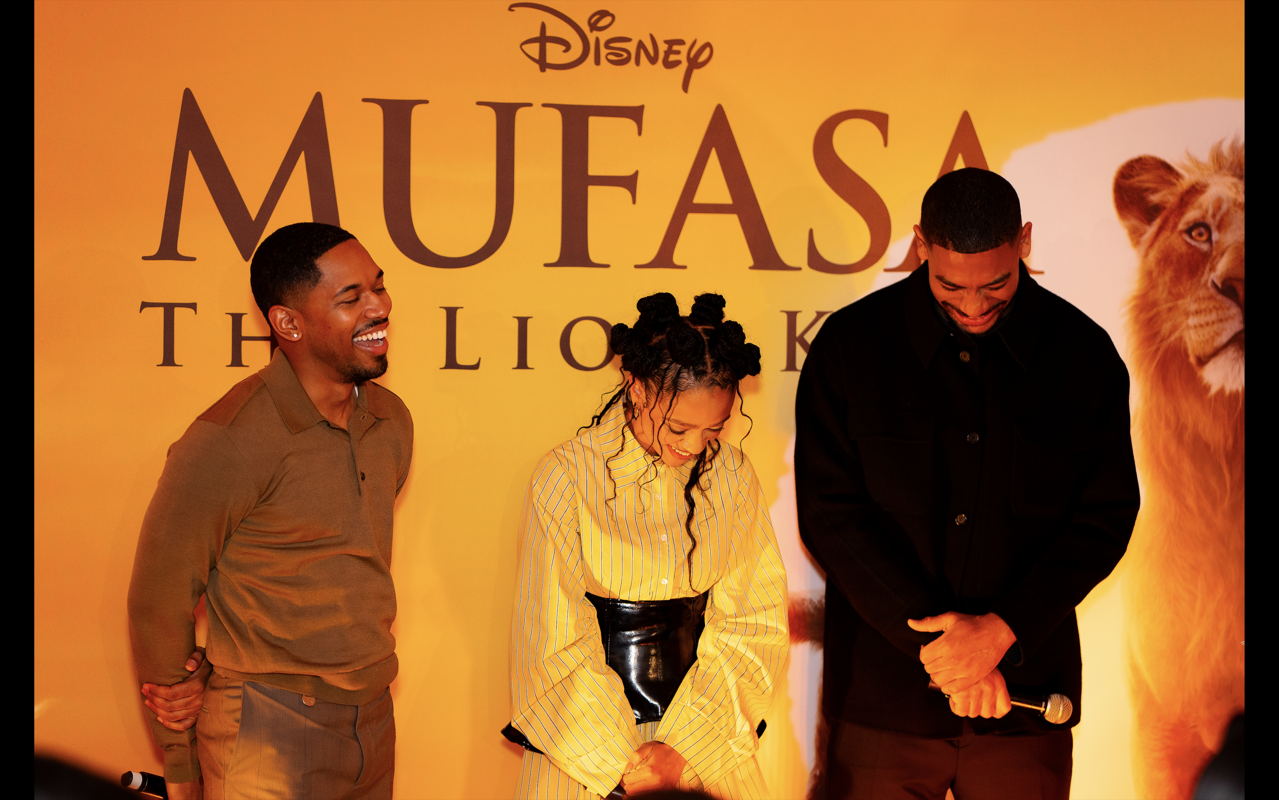 Three people are smiling and laughing in front of a yellow backdrop with the text 'Disney The Lion King Mufasa.' One person is a woman with braided hair wearing a yellow striped shirt; the other two are men, one wearing a brown shirt and the other wearing a black shirt, both with short hair. They are interacting and appear to be enjoying the moment.