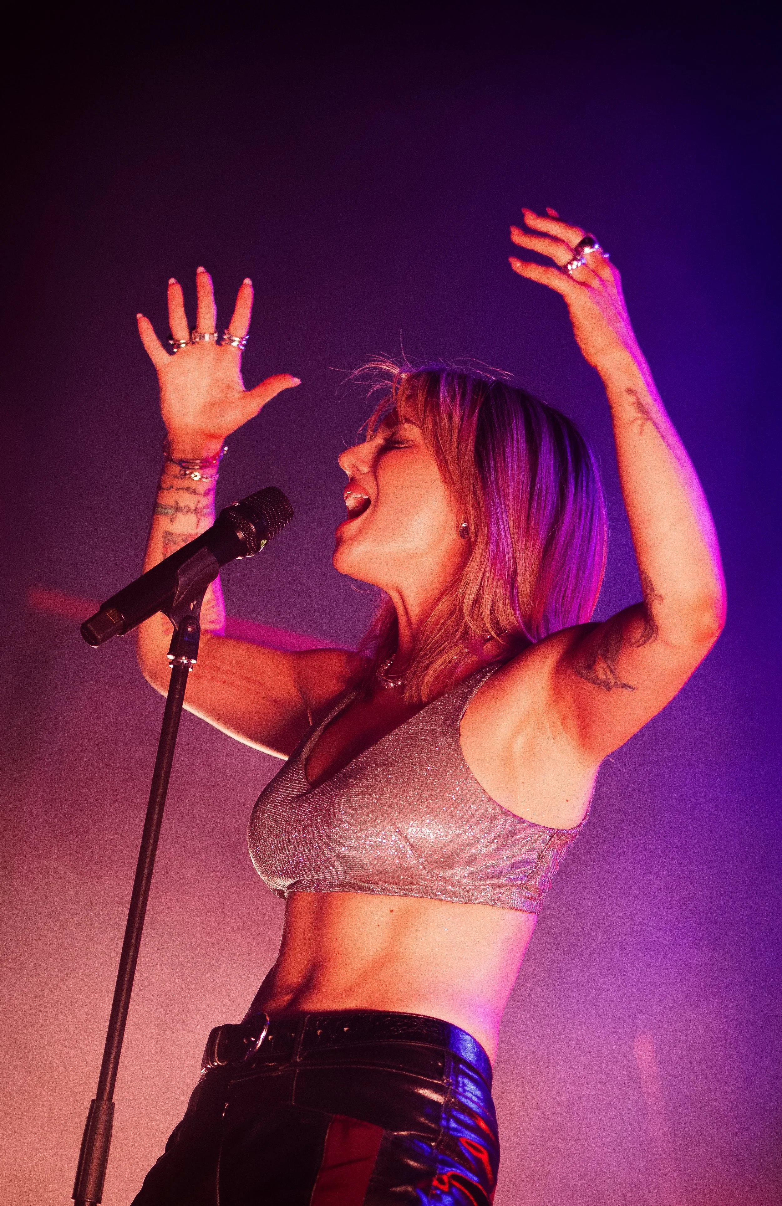 A woman singing into a microphone on stage, raising her hands with rings and tattoos, wearing a sparkling crop top and high-waisted pants, with colorful lighting creating a vibrant atmosphere.