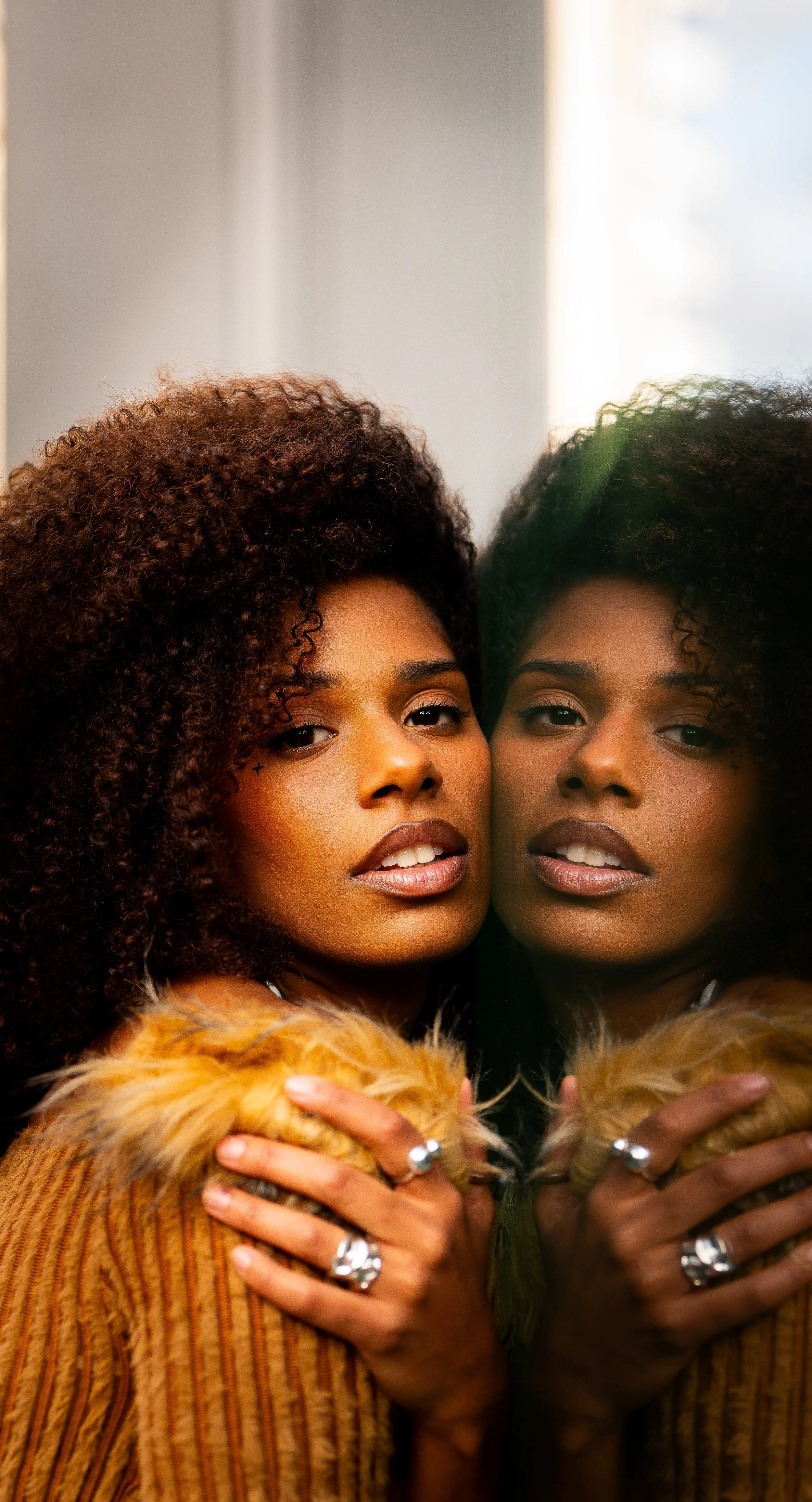 A woman with curly hair looking into a mirror, with her reflection visible. She is wearing a fur collar and has jewelry on her fingers.