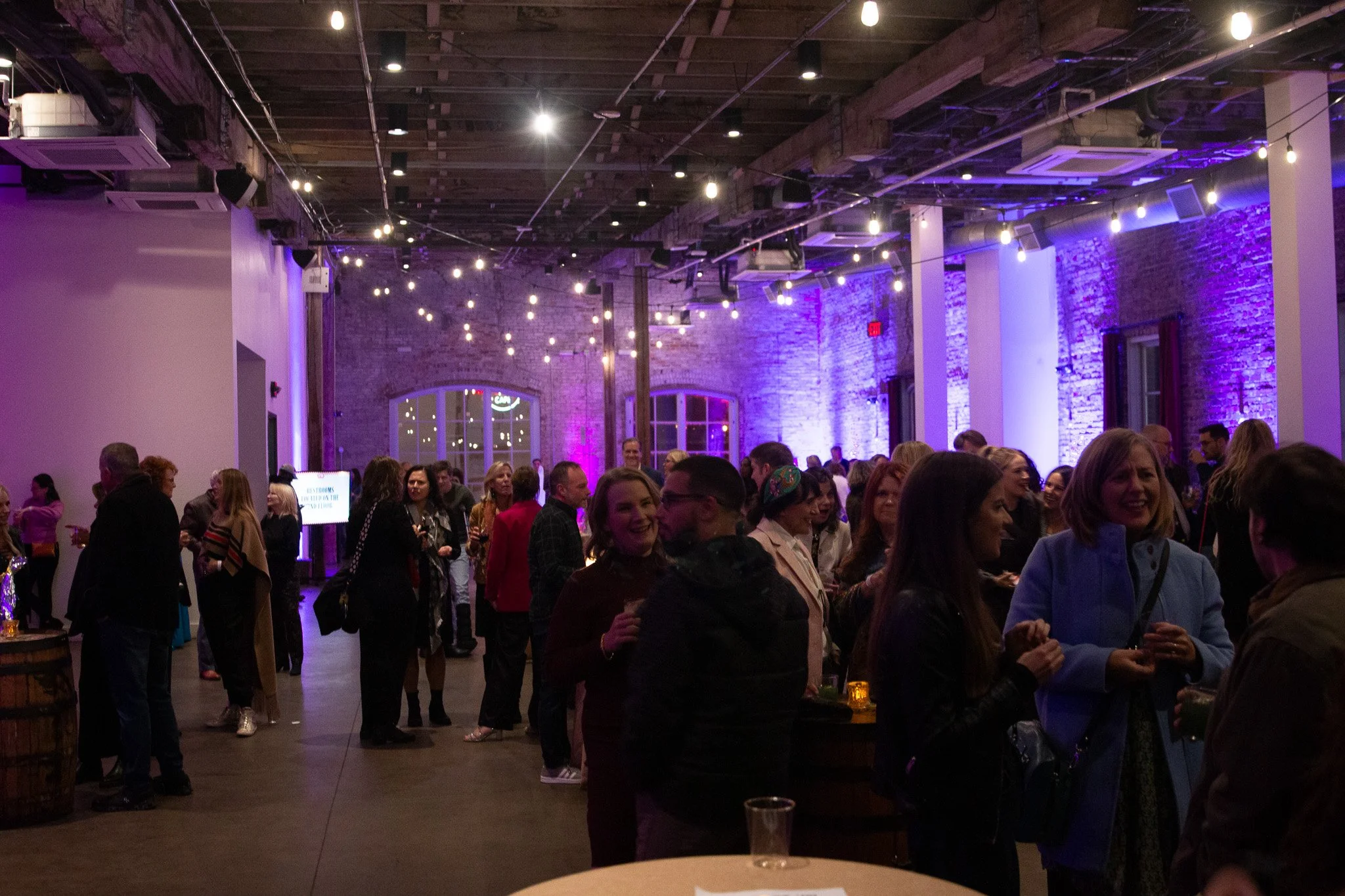 A crowded indoor event (A Dolly Good Time) with people socializing and smiling, illuminated by string lights and purple accent lighting on exposed brick walls.