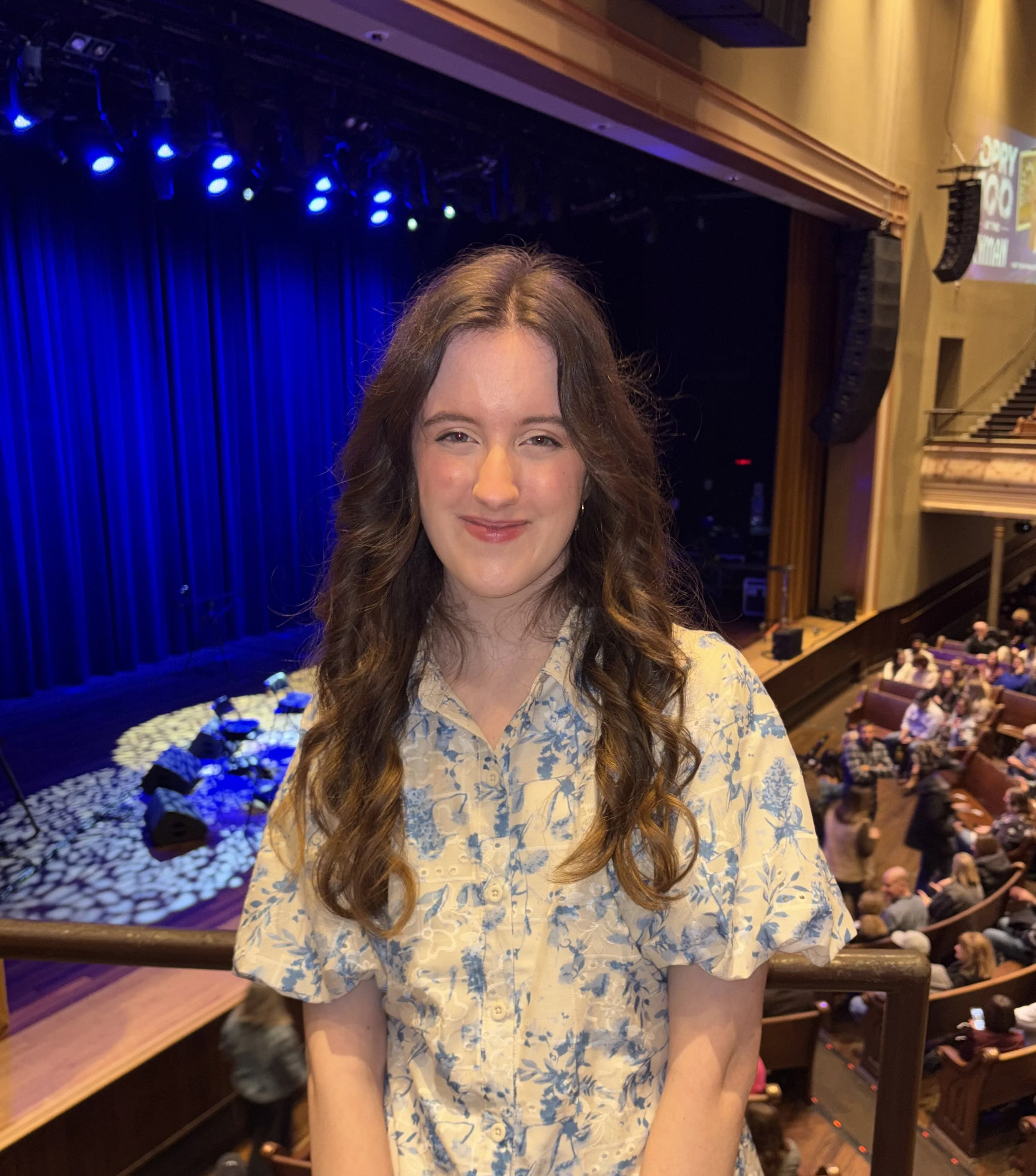 A smiling woman with long wavy hair wearing a yellow floral dress at a theater, with a stage with blue curtains and blue stage lights in the background, and an audience seated in front of the stage.