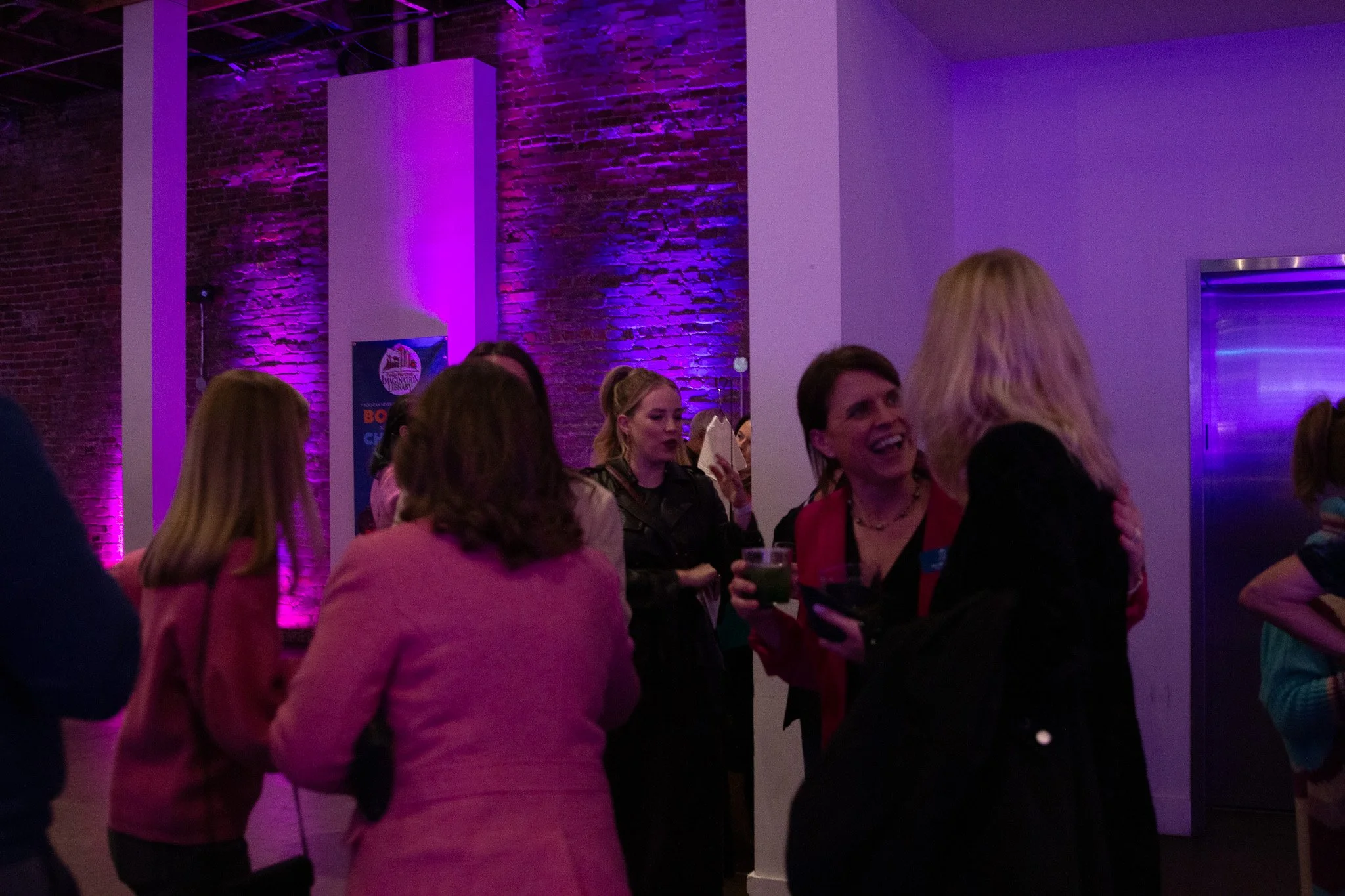 People socializing at an indoor event with purple and pink lighting, brick wall backdrop, and a poster on the wall. A Dolly Good Time.