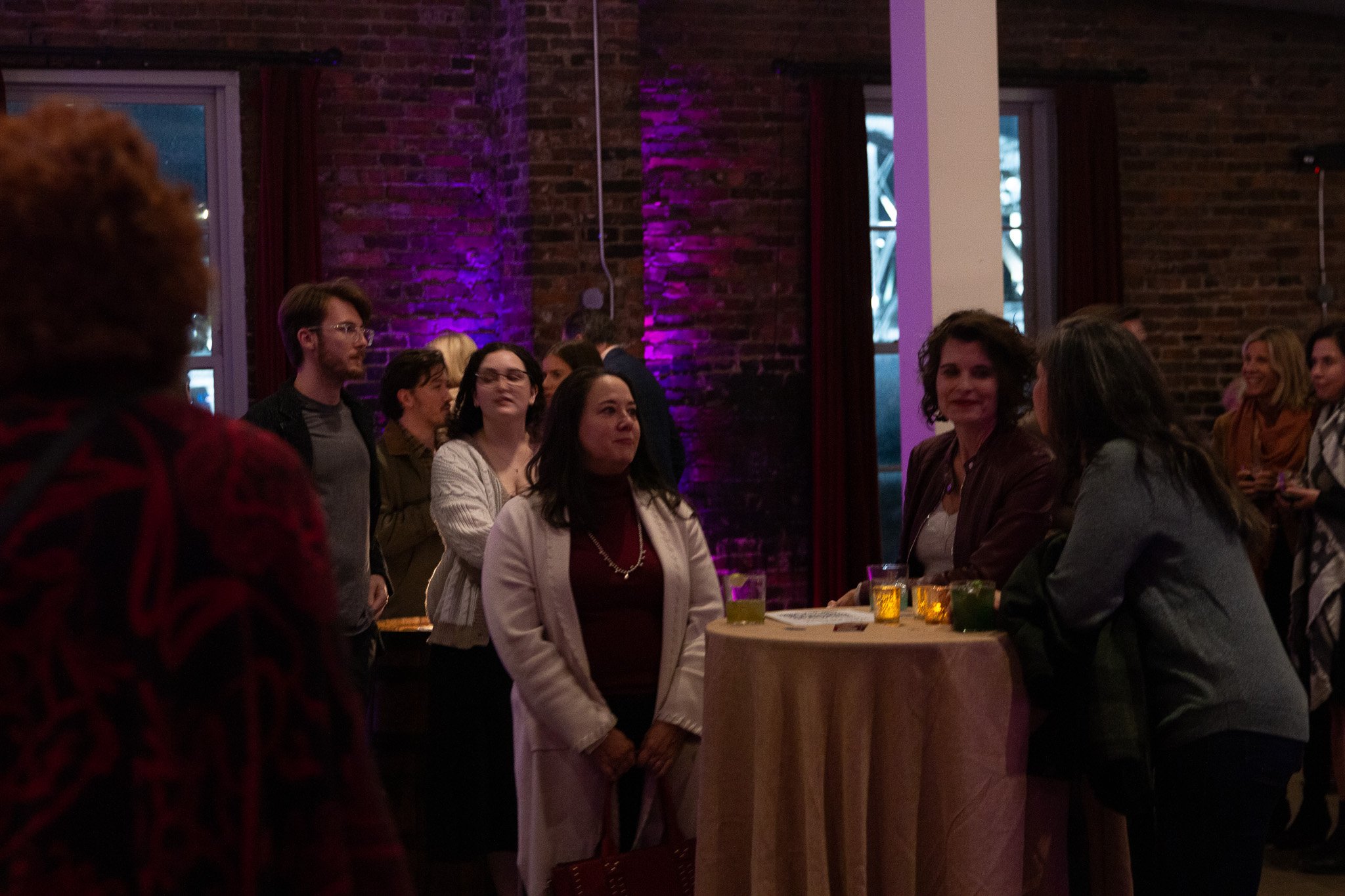 People gathered at a A Dolly Good Time event in the past in a dimly lit room with brick walls and purple lighting, some standing around a high table with drinks.