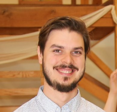 Portrait of a smiling young man with dark hair and a beard, wearing a light blue shirt, in front of a wooden interior.