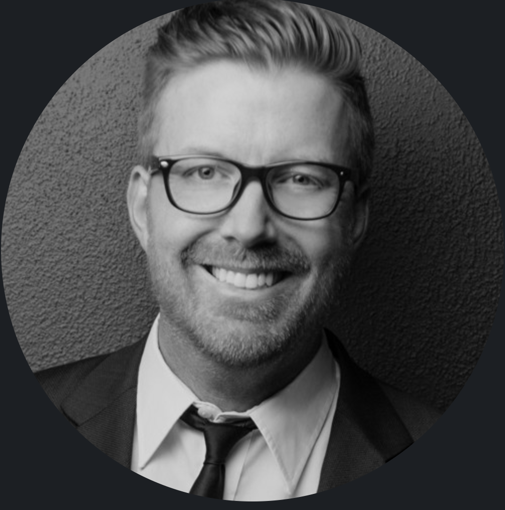 Black and white portrait of a smiling man with glasses, short hair, and a beard, wearing a suit and tie.