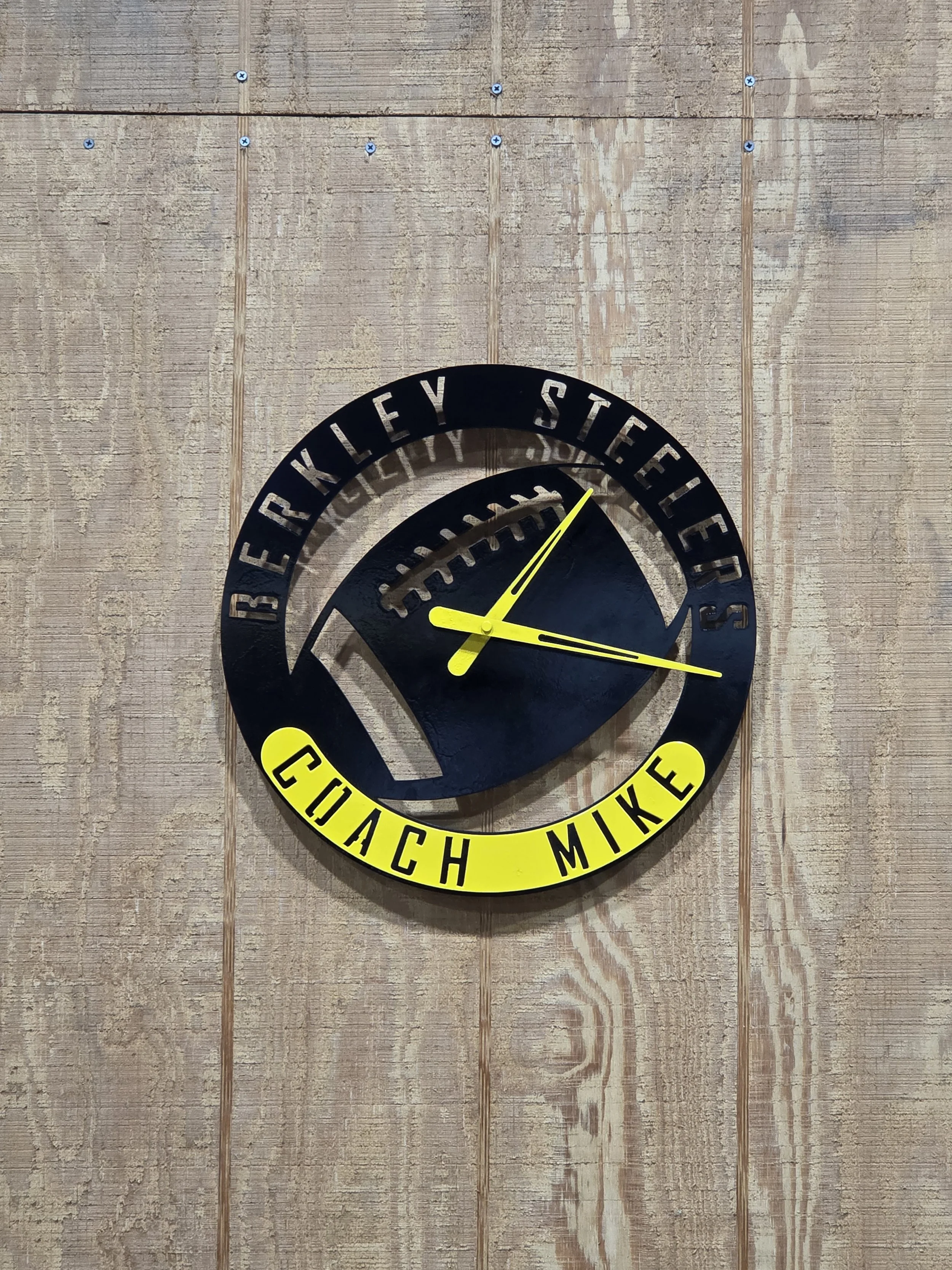 A wall clock with a black face and yellow hands, mounted on a wooden wall. The clock has a yellow ring around it with the words 'COACH MIKE' in black lettering, and the black face has the phrase 'REALLY STEELER' in there.