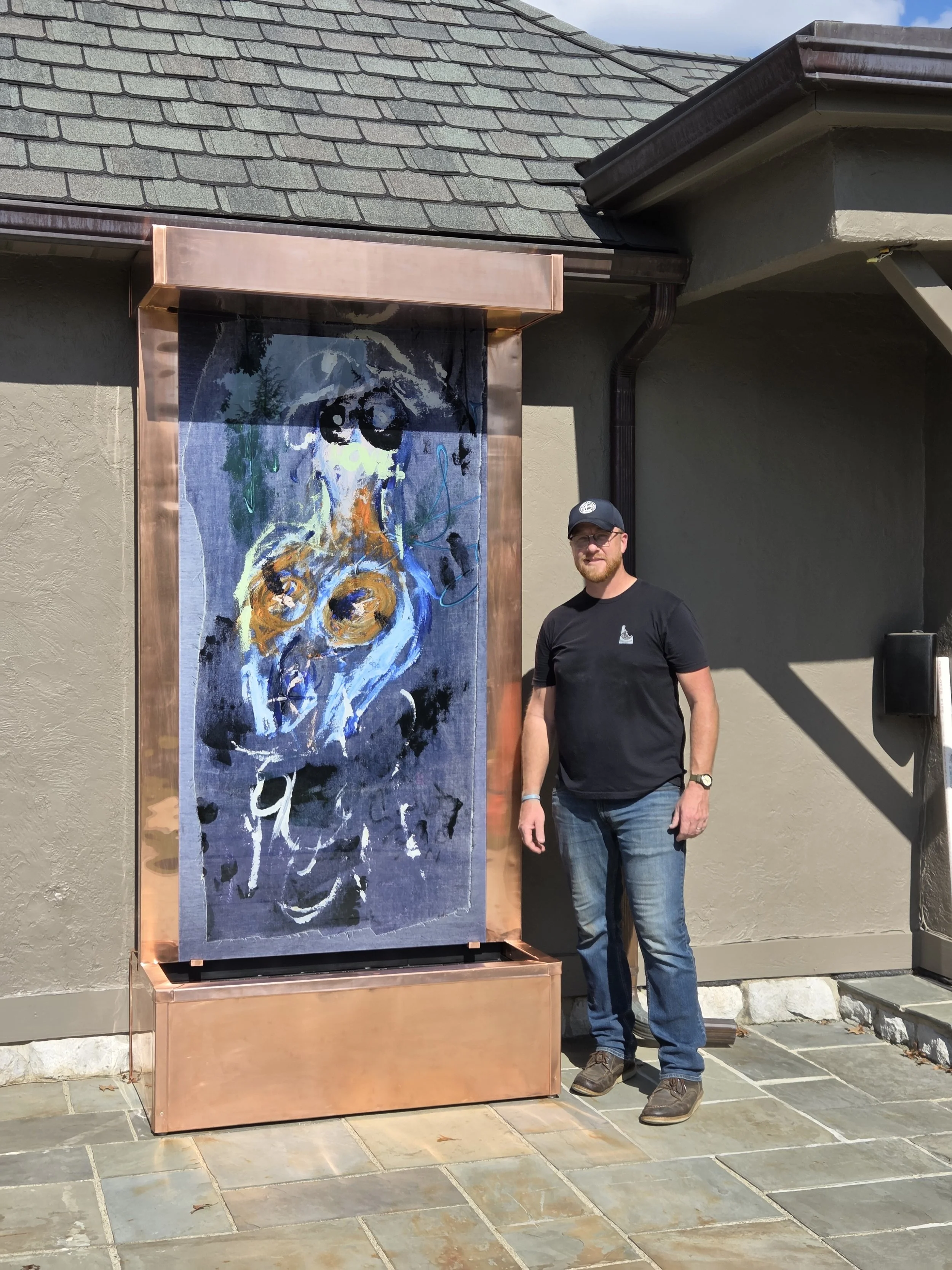A man wearing a black t-shirt, jeans, and a cap stands next to a large, colorful abstract painting displayed outdoors on a copper frame. The background shows a building with a tiled roof and a beige wall, with sunlight casting shadows.