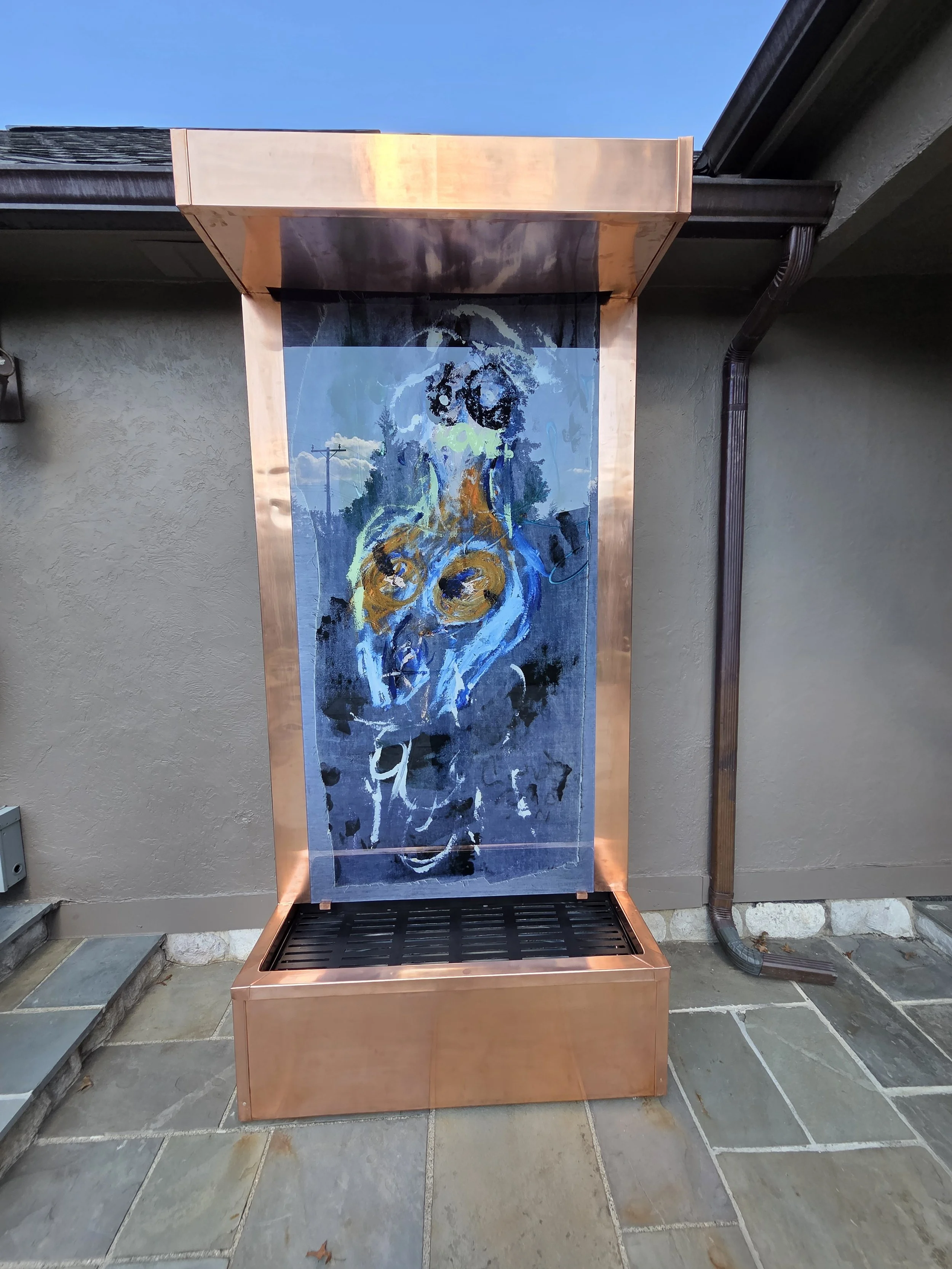 Outdoor copper and glass water feature with artistic image of a dog etched on the glass, set against a beige stucco wall and stone paving.