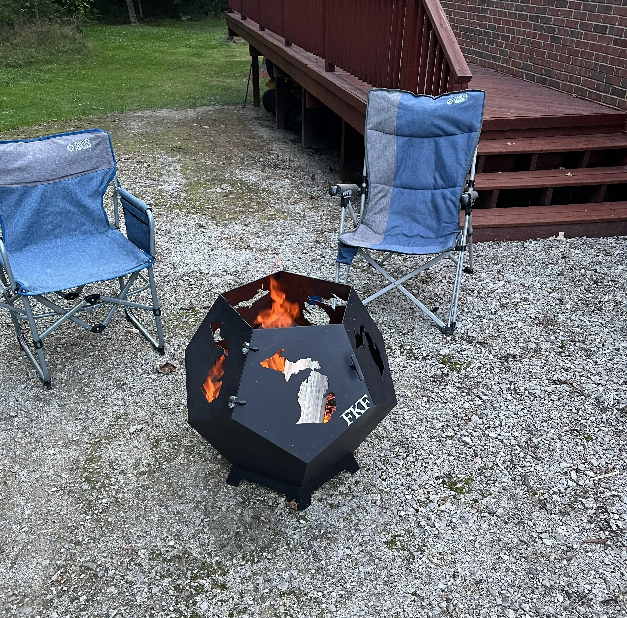A backyard scene with a red wooden deck and stairs, two blue foldable chairs, a small fire pit with a fire burning, and grass area in the background.
