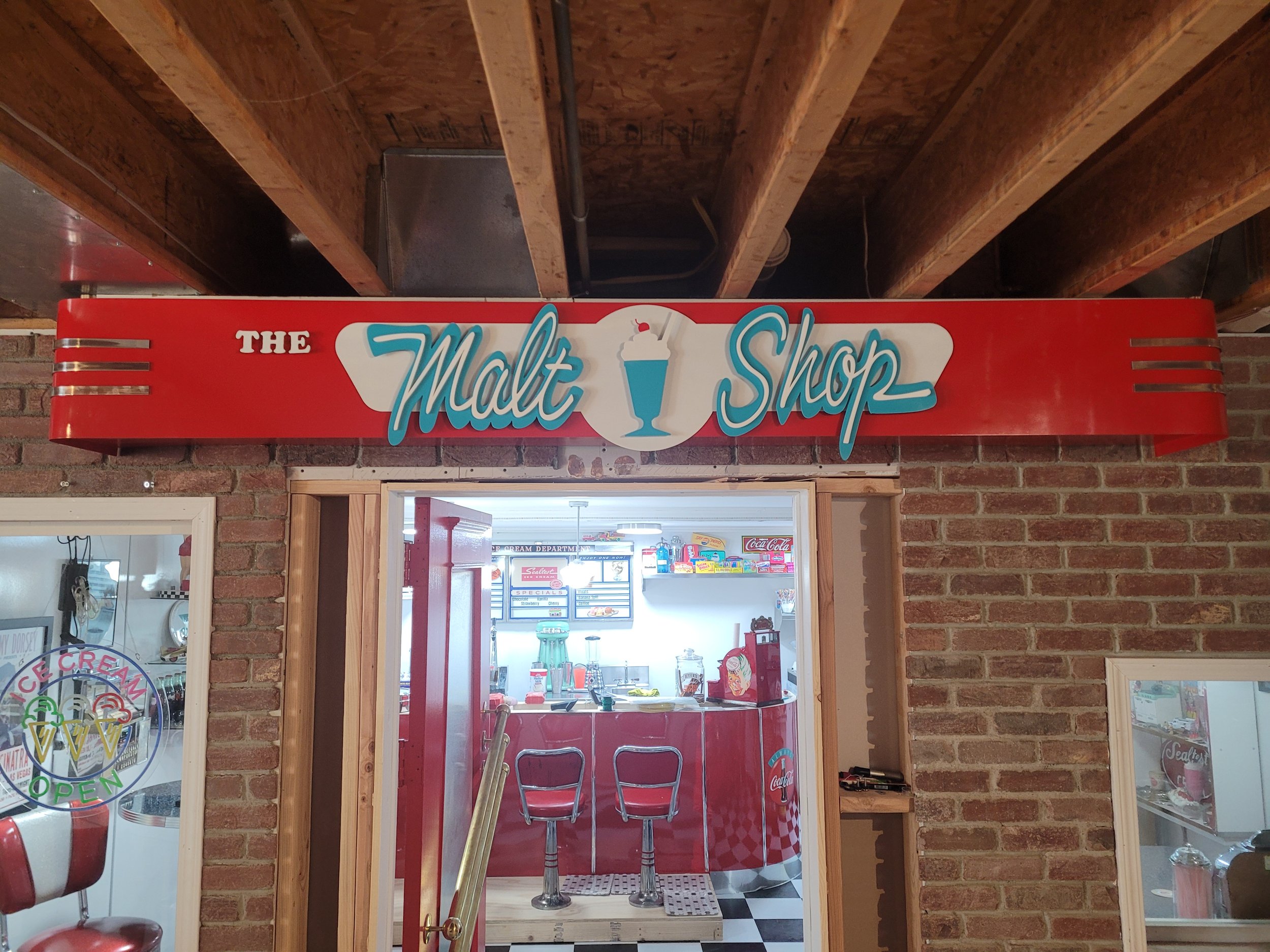 Interior view of an ice cream shop with a bright red sign reading 'The Malt Shop' hanging above the entrance. The shop features brick walls, a counter with red and white checkered patterns, and vintage decor. Visible through the window are various ice cream and soda fountain items.