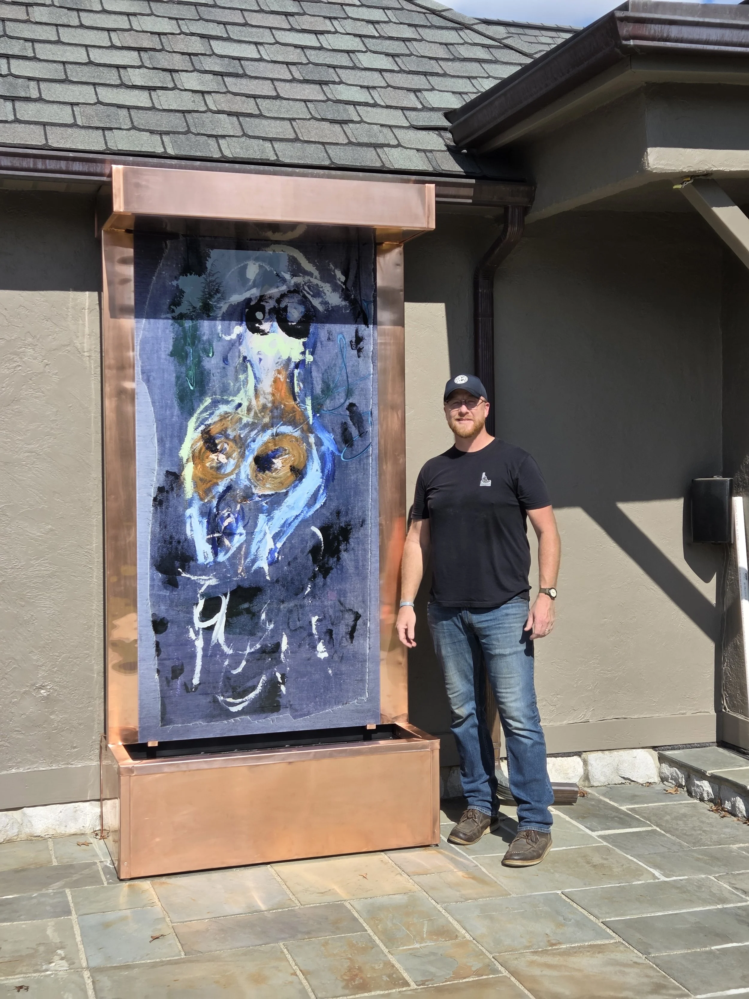 A man with glasses, a black t-shirt, jeans, and a cap, standing outside next to a large abstract painting on a copper frame attached to a building wall.