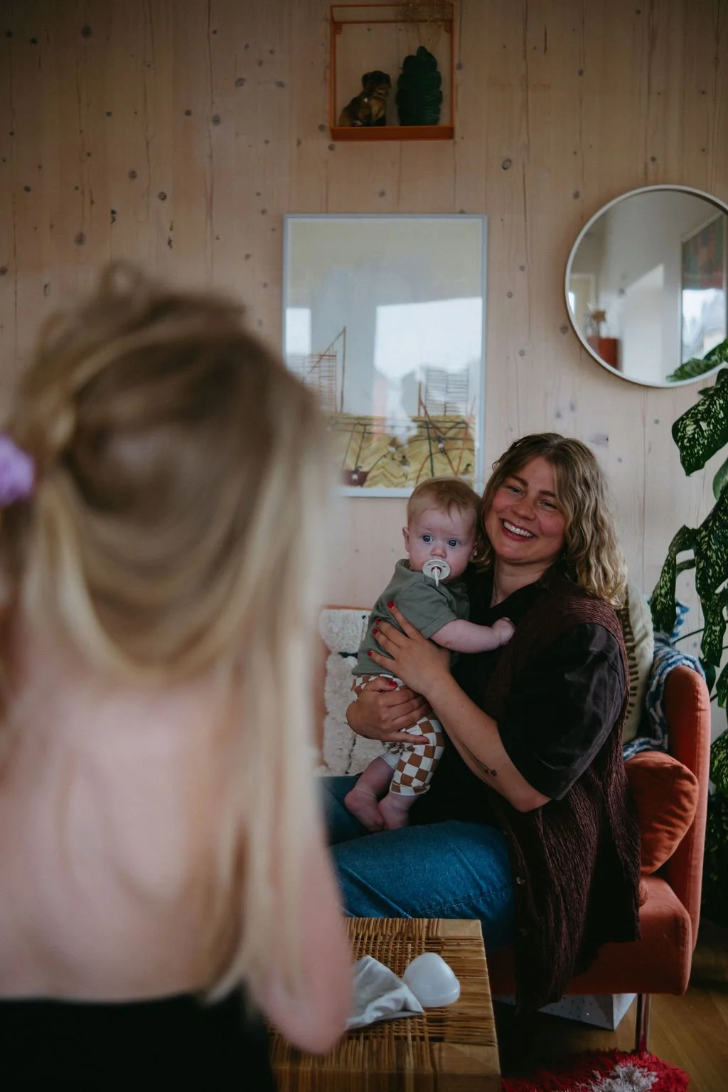 En kvinna sitter i ett vardagsrum och håller i en bebis med blont hår och pacifier i munnen, kvinnan ler glatt. En person med blont hår, troligen ett barn, är i förgrunden med ryggen mot kameran, något utanför fokus.