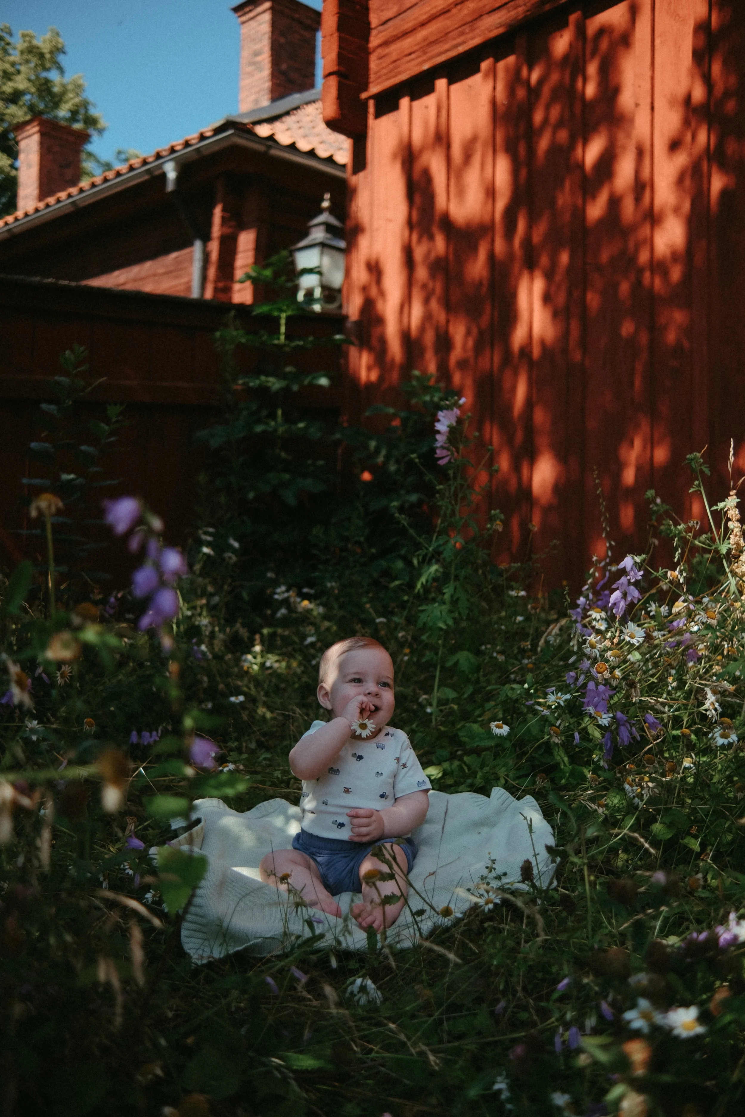 En glad liten bebis som sitter på en filt bland blommor i en trädgård med röda husväggar i bakgrunden.