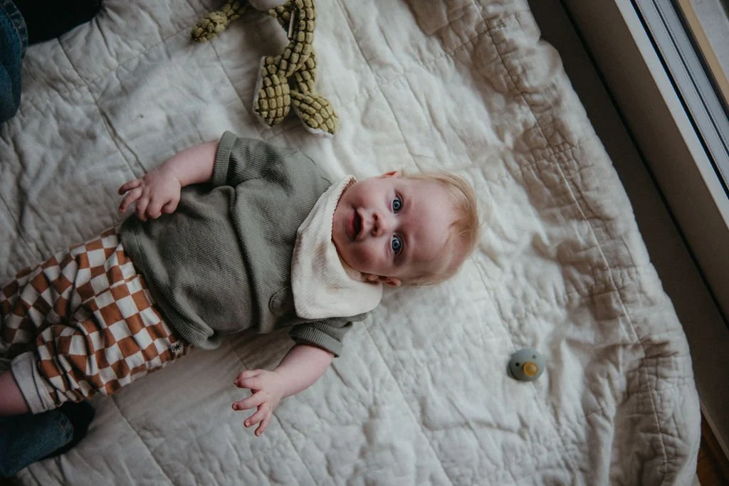En baby ligger på en vit quilt på golvet, omgiven av leksaker inklusive en krokodil och en liten figur.