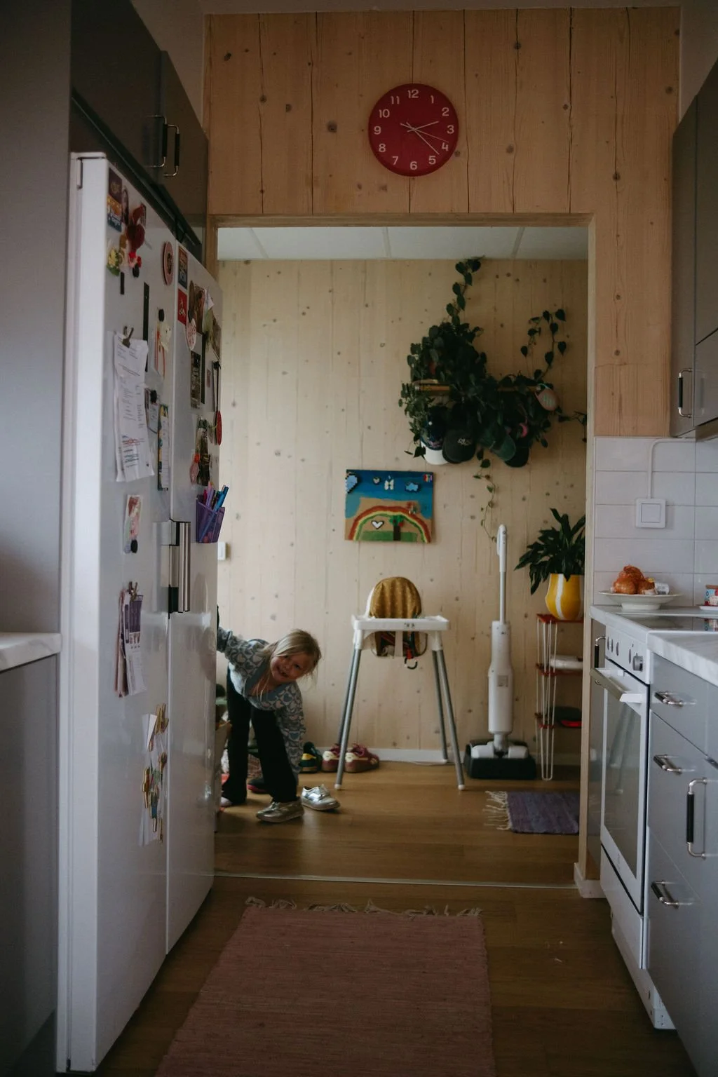 Kök med barn som leker och klär på sig, magneter på kylskåpet, väggklocka, växter, tavla och köksutrustning