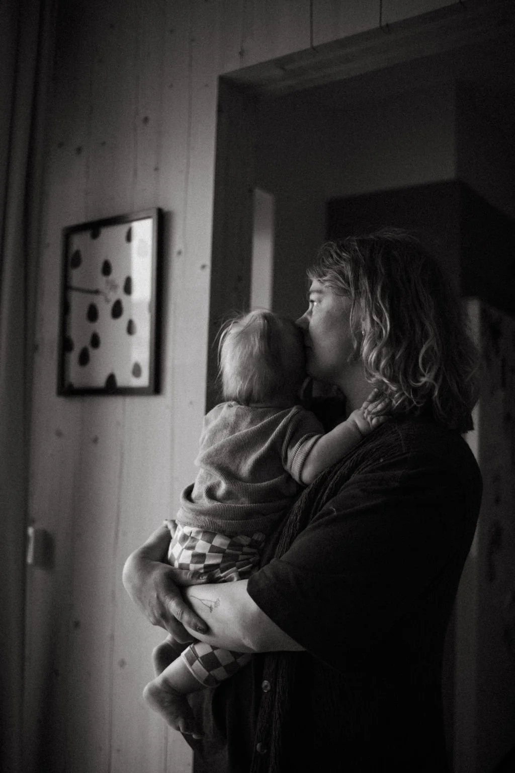 En kvinna håller ett barn och kysser dem på kinden i ett inomhus. Svartvitt foto.